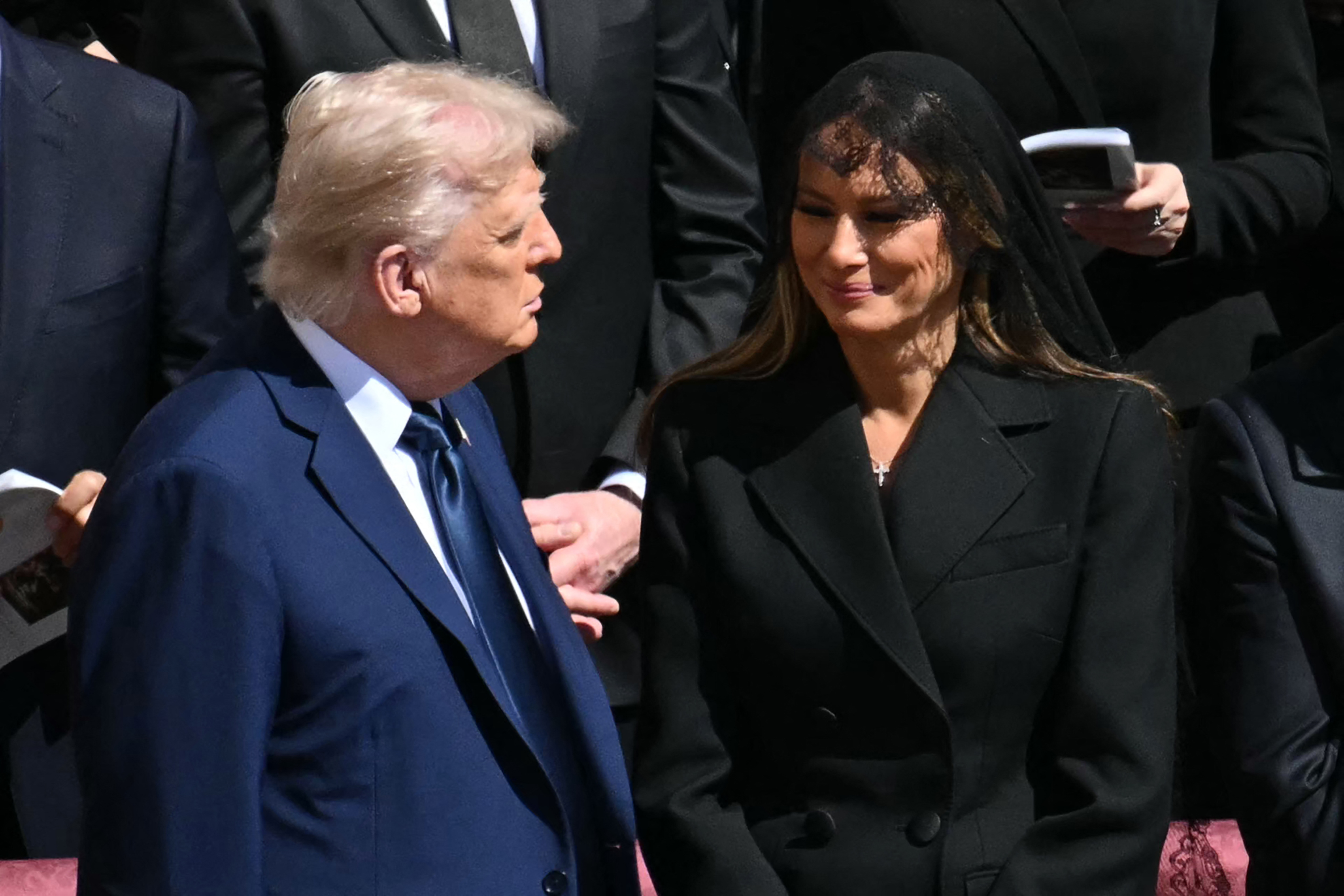Donald und Melania stehen zusammen und unterhalten sich. Melania lächelt sanft, als sie inmitten der Trauer einen Moment der stillen Freude teilen.