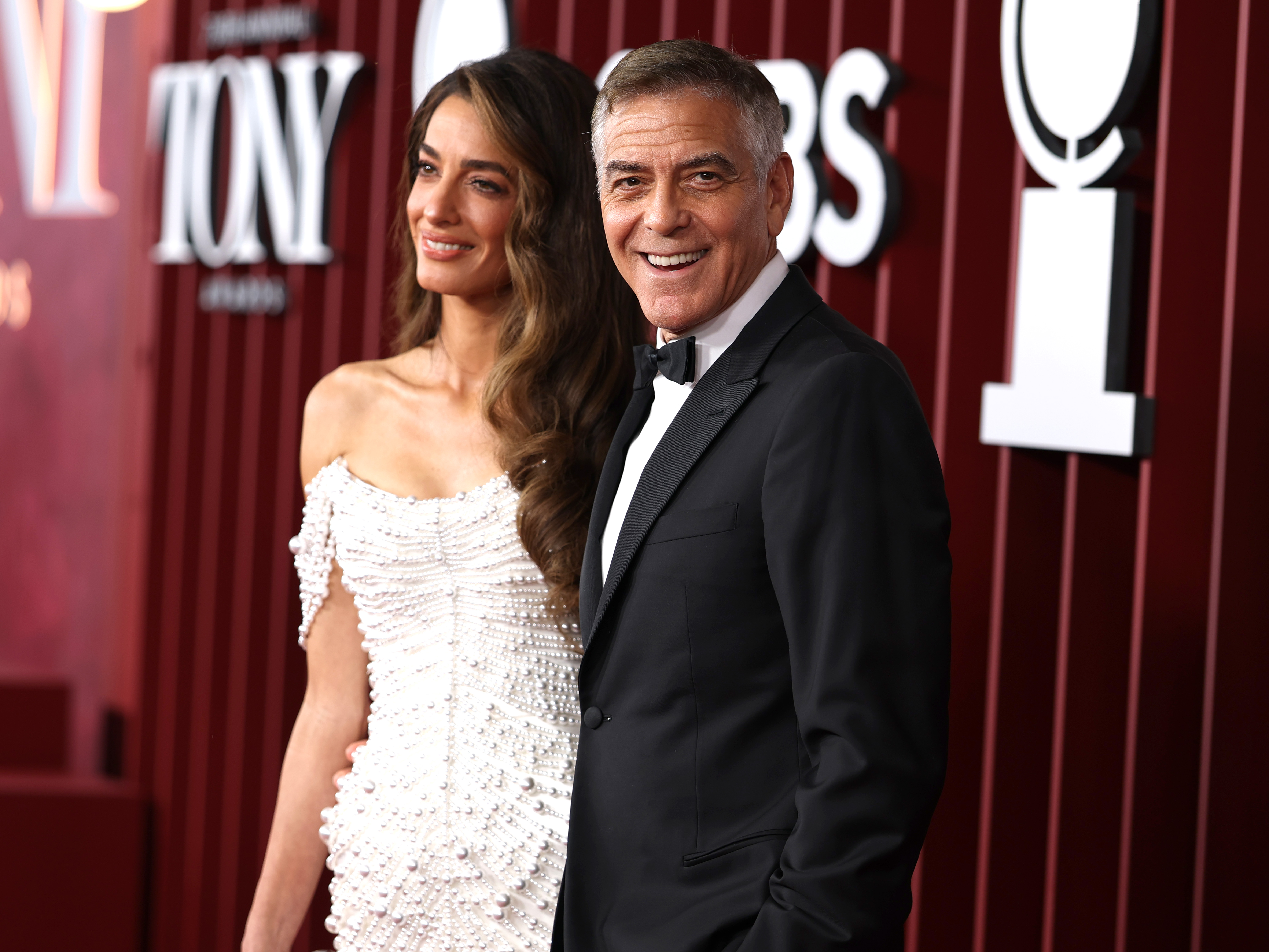 George Clooney lächelt sanft in die Kameras, während Amal Clooney neben ihm steht. Ihr perlenbesetztes Kleid glitzert im Scheinwerferlicht, als sie gemeinsam auf dem roten Teppich der Tony Awards posieren.