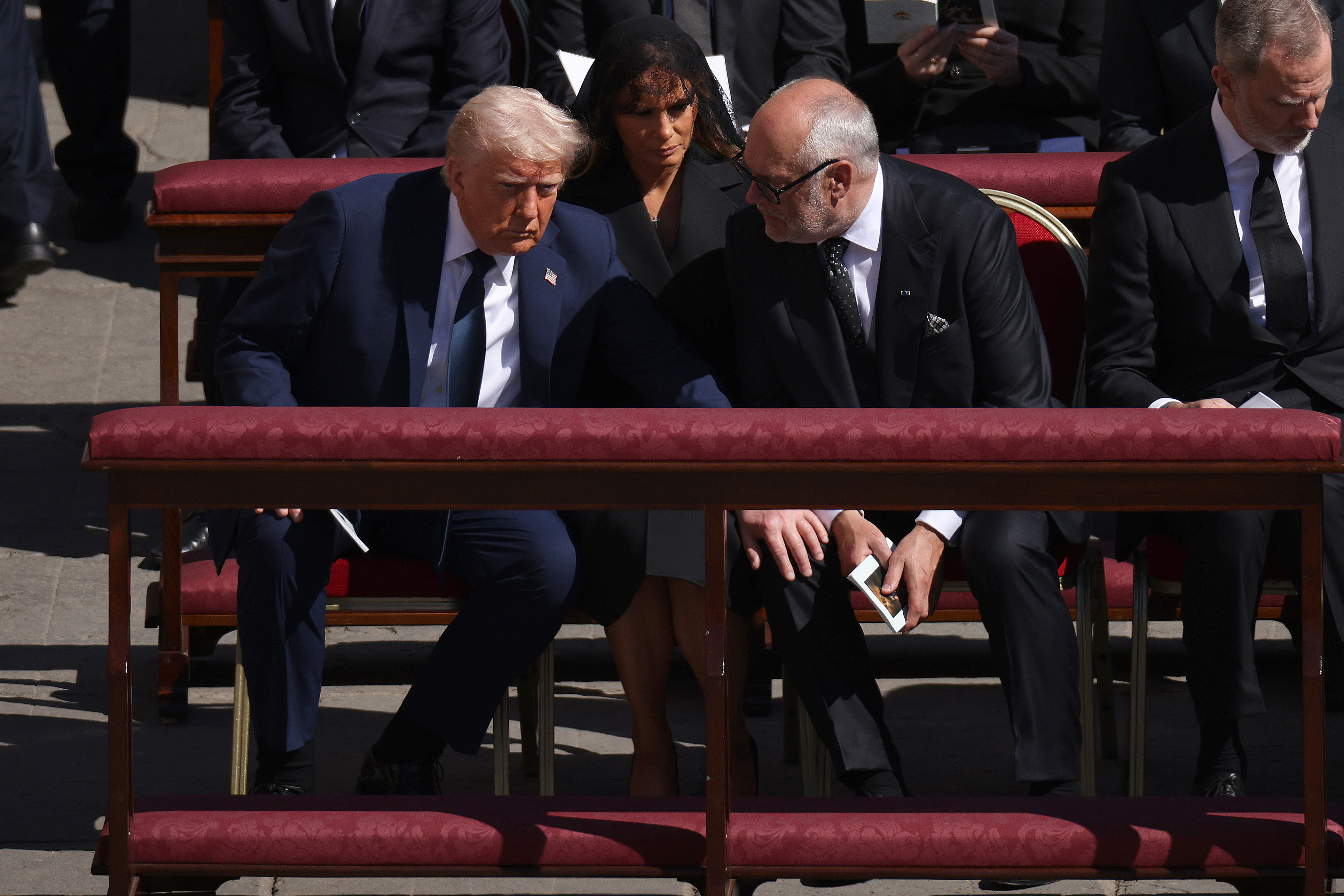 Donald und der estnische Präsident Alar Karis beugen sich während der Beerdigung in einem ruhigen Gespräch vor. Währenddessen sitzt Melania ruhig und gelassen da und beobachtet das Geschehen.