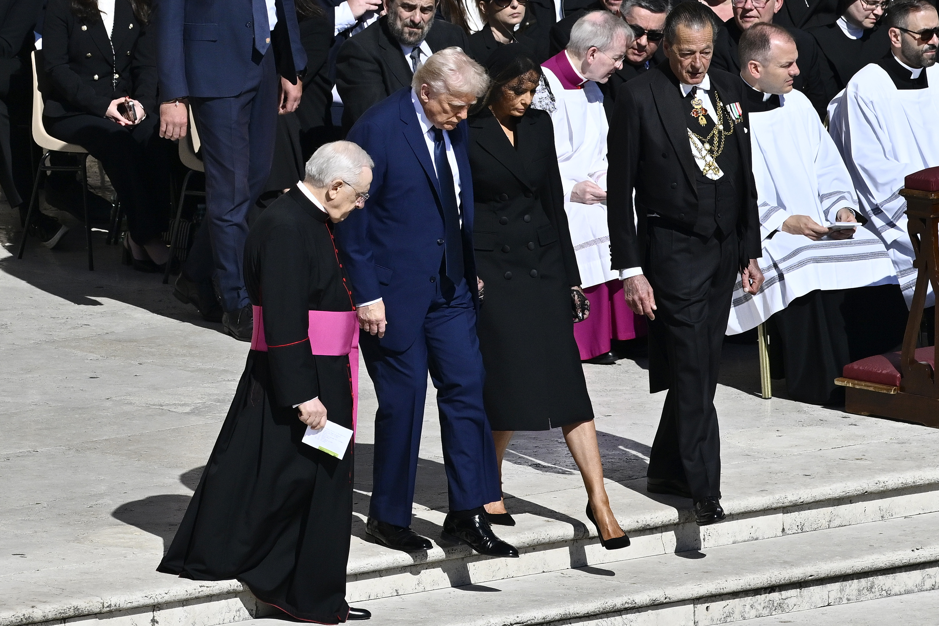 Donald und Melania Trump betreten den Petersplatz, begleitet von einem katholischen Priester. Während der Priester in ein schwarzes Gewand gekleidet ist, wie auch Melania in ihrem Outfit, hebt sich Donalds blauer Anzug in einem starken Kontrast ab und zieht die Aufmerksamkeit auf sich inmitten des Meeres dunkler Kleidung.