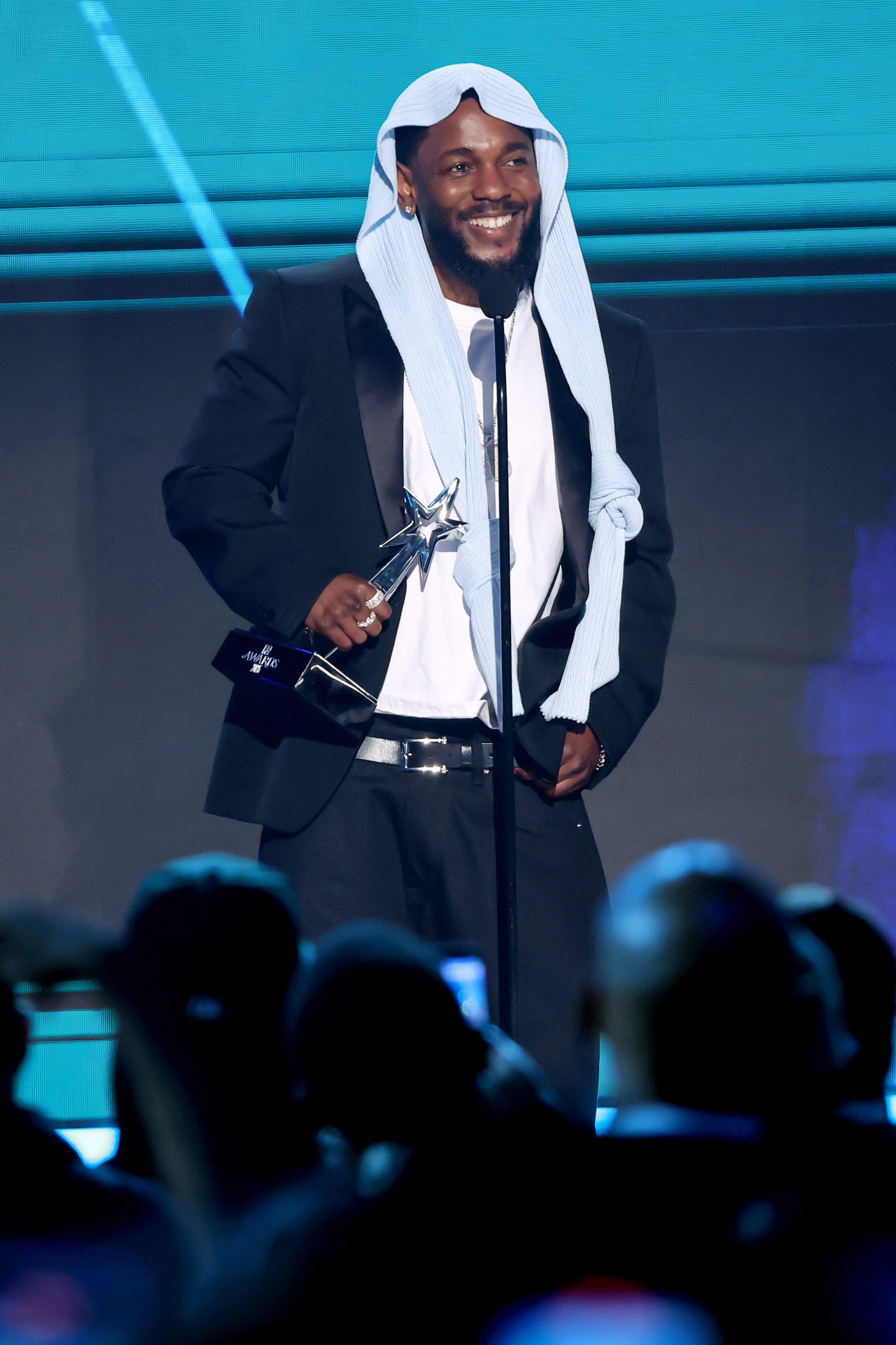 Kendrick Lamar zeigt ein strahlendes Lächeln, während er bei den BET Awards 2025 auf der Bühne eine Trophäe hält. Mit seiner mittlerweile ikonischen Kopfbedeckung und seinem schwarzen Anzug schien der Rapper wirklich gerührt über die anhaltende Anerkennung des Abends.