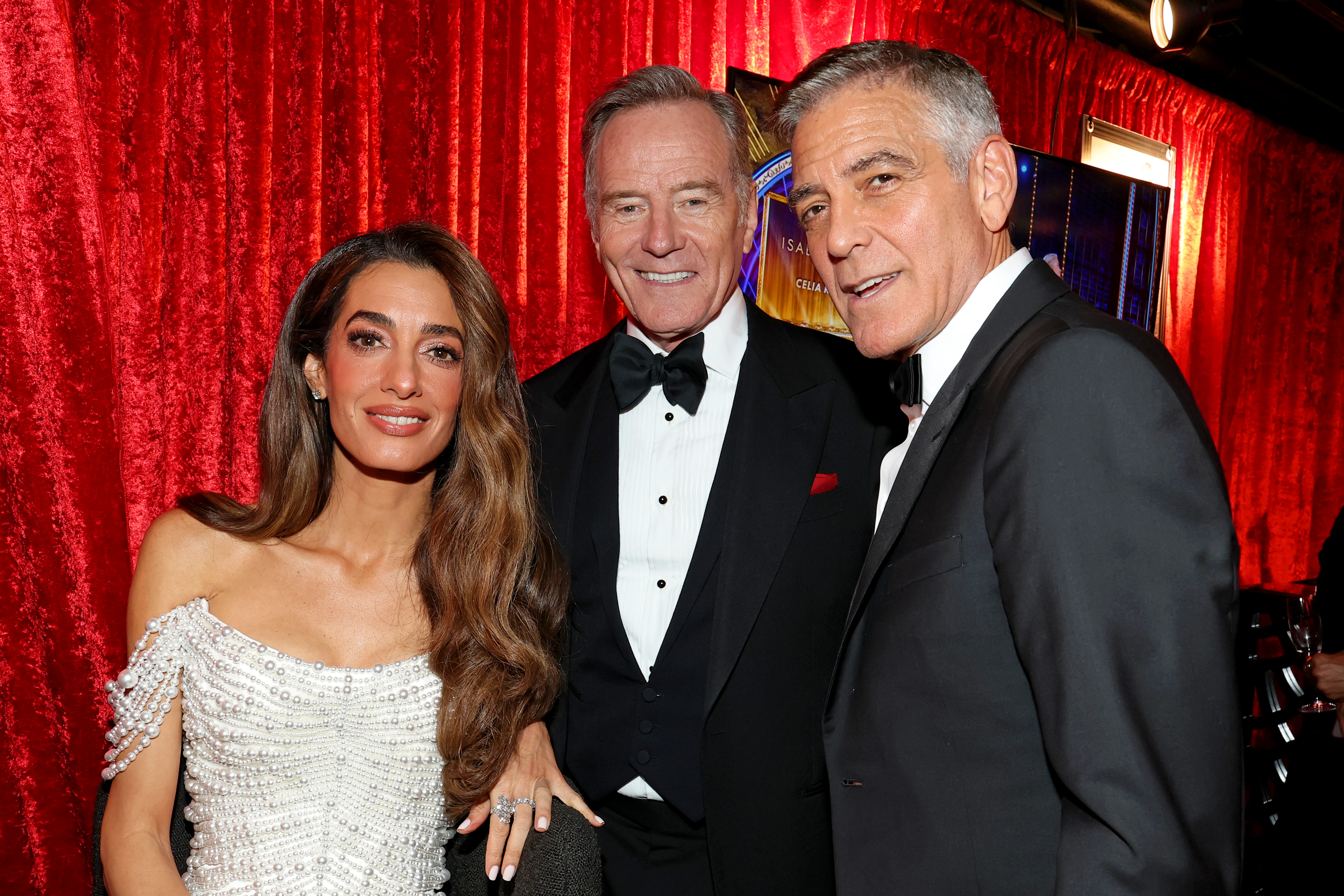 Amal und George Clooney treffen Bryan Cranston hinter der Bühne im Green Room. Eingerahmt von roten Samtvorhängen lächelt Amal direkt in die Kamera, während George sich ihr mit einem entspannten Gesichtsausdruck zuneigt.