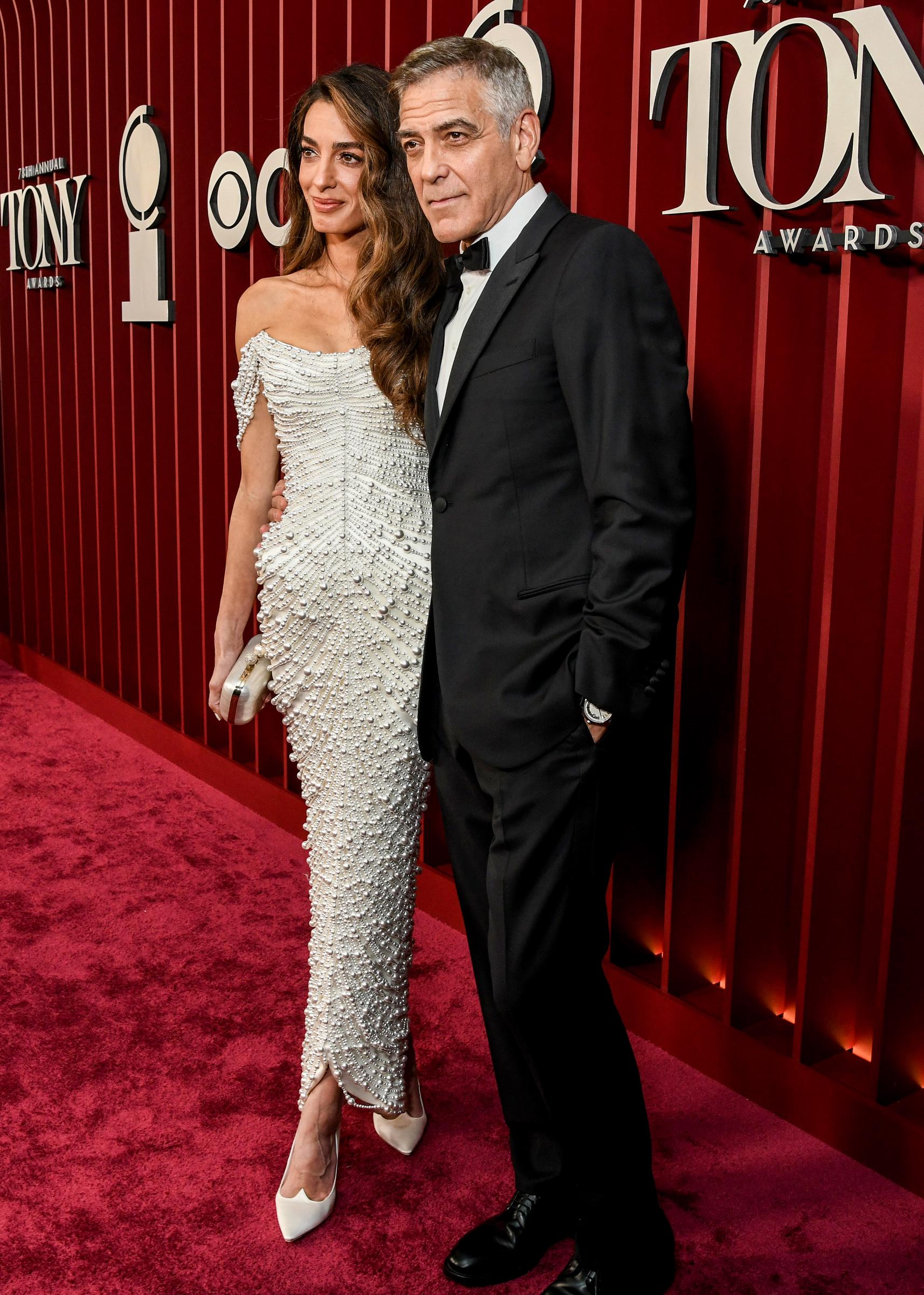 Amal Clooney steht mit gekreuzten Füßen neben George Clooney bei den Tony Awards. George wirkt entspannt, als sie gemeinsam vor der tiefroten Kulisse posieren.