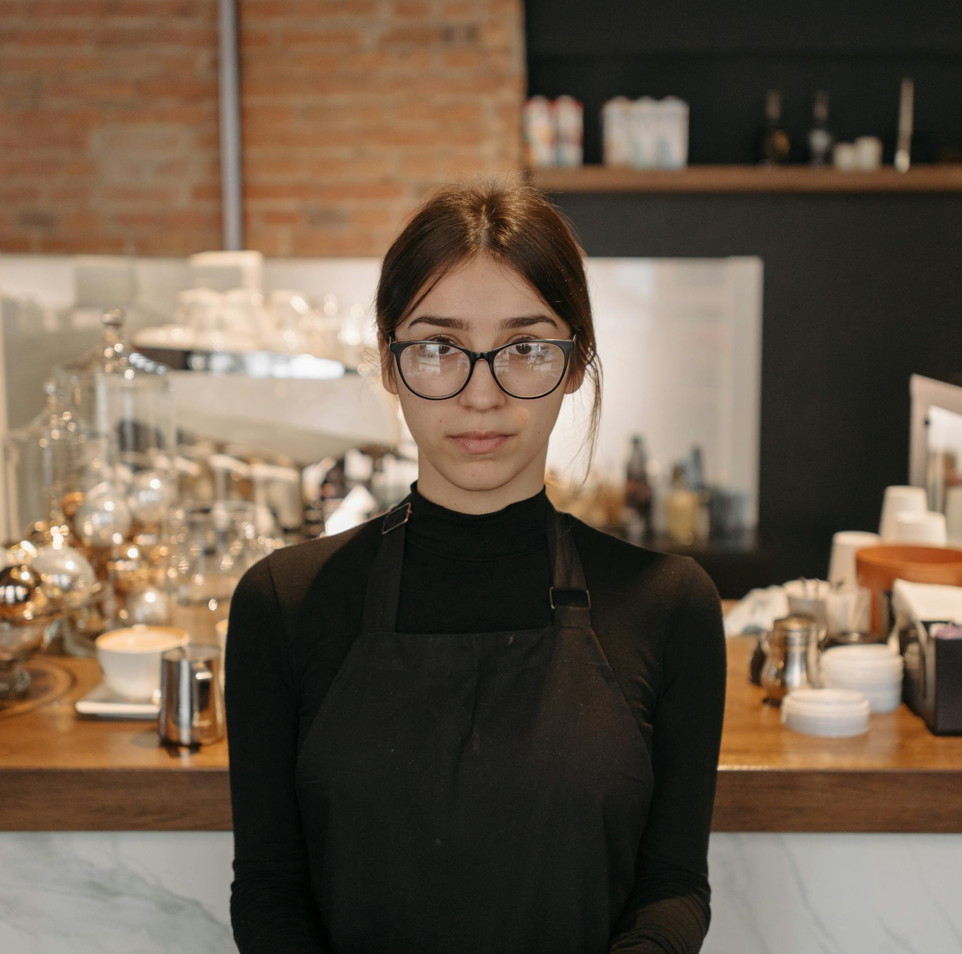 Eine Kellnerin mit Brille und Schürze steht hinter einem Tresen in einem Café | Quelle: Pexels