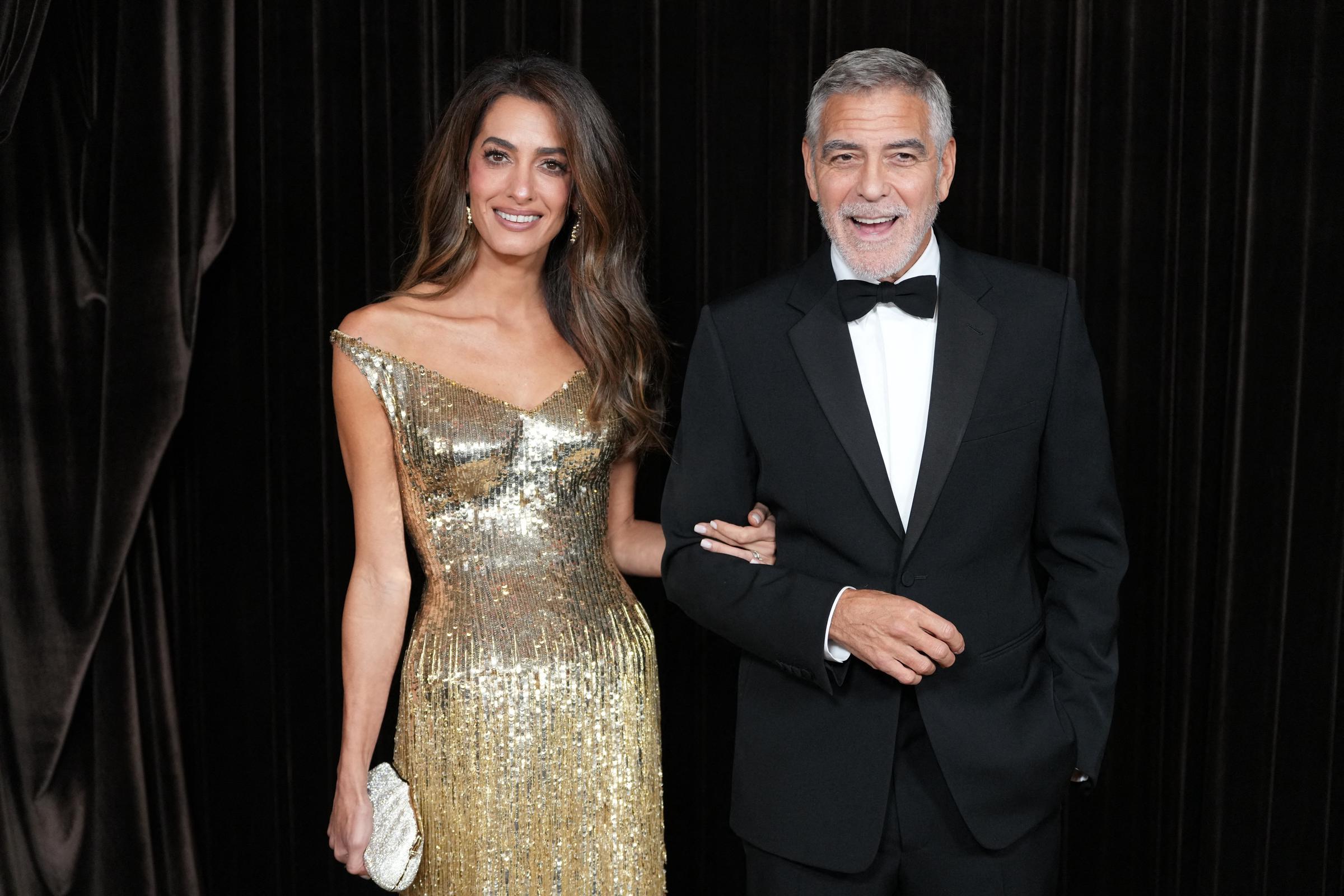 George und Amal Clooney posieren Arm in Arm bei den Albies 2026 am 9. Oktober in London. Amal strahlt in einem schimmernden, schulterfreien Goldkleid mit passender Clutch, während George einen klassischen schwarzen Smoking mit Fliege trägt. | Quelle: Getty Images