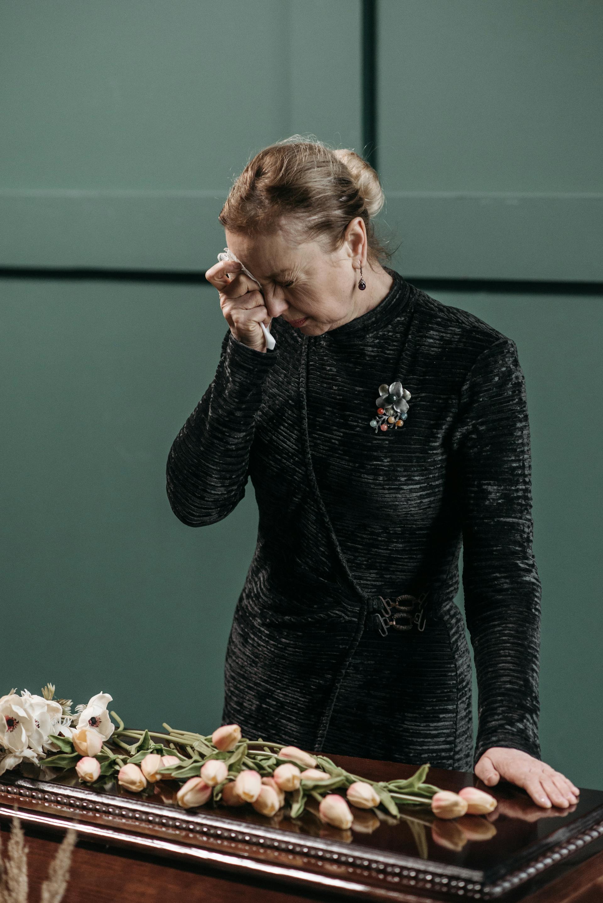 Eine weinende reife Frau in einem schwarzen Kleid, die sich eine Träne mit einem Taschentuch abwischt, während sie neben einem Sarg steht | Quelle: Pexels