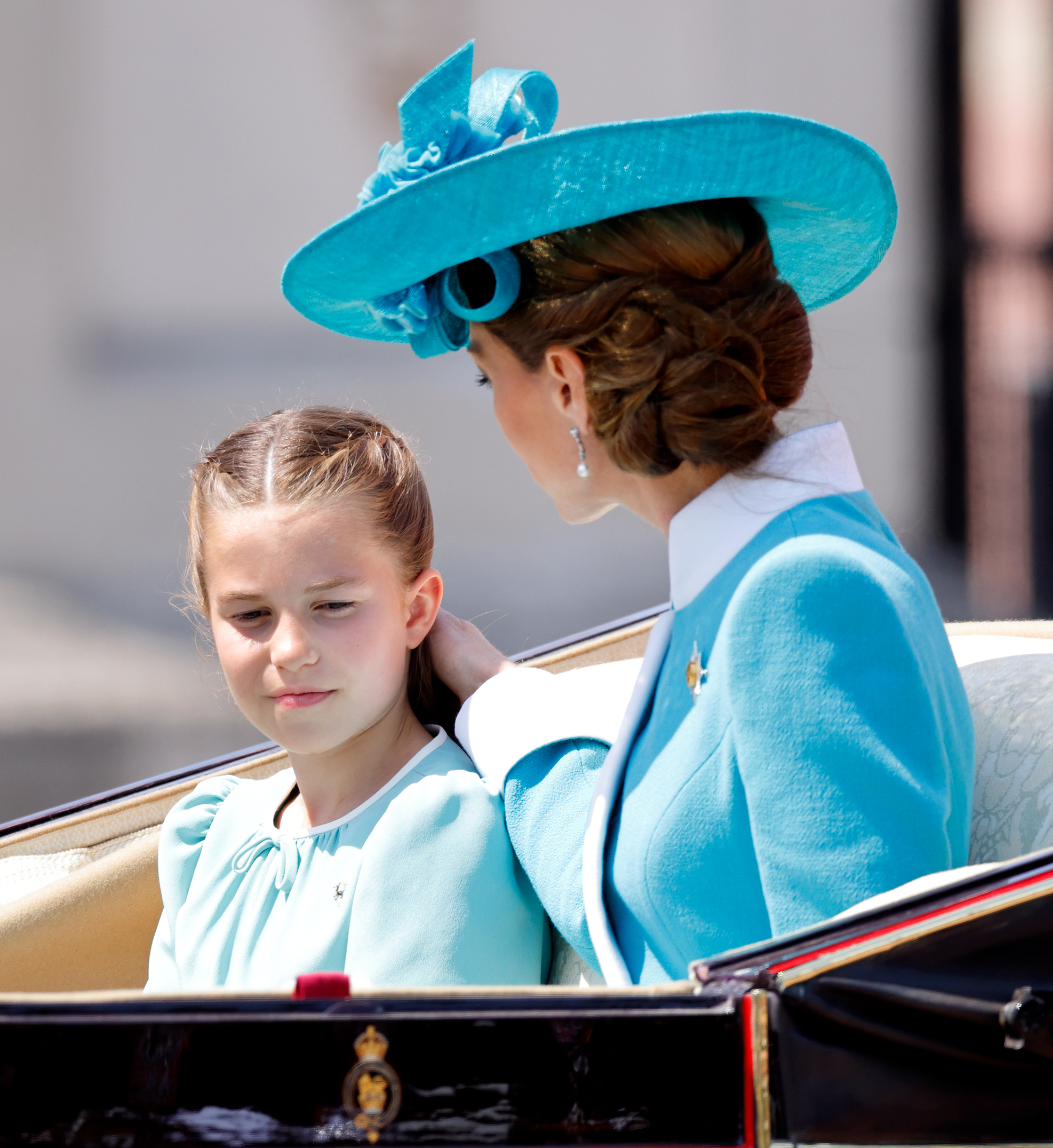 Prinzessin Charlotte von Wales und Catherine, Prinzessin von Wales, kehren in den Buckingham Palace zurück. Catherine strahlt in ihrem aquamarinfarbenen Kleid, dem breitkrempigen Hut und den brünetten Locken, die zu einer weichen Hochsteckfrisur gestylt sind, während Charlotte in ihrem weiß-aquamarinfarbenen Kleid und ihrem voluminösen braunen Haar, das locker mit einem ordentlichen Mittelscheitel getragen wird, glänzt.