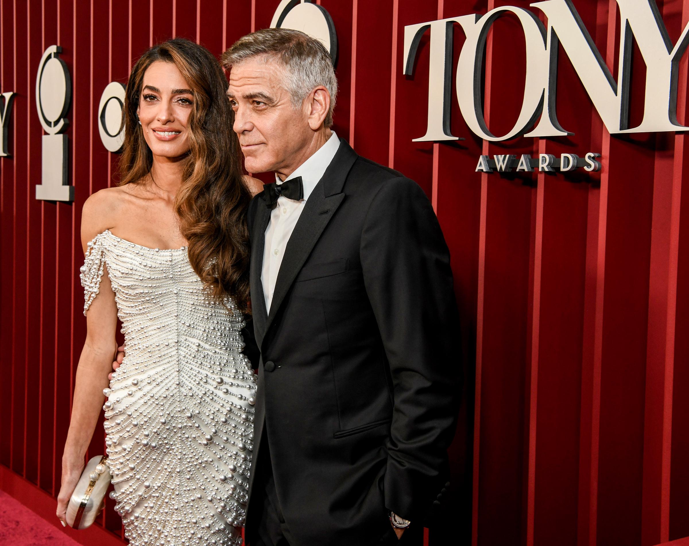 Amal Clooney lächelt sanft, ihre perlenbesetzte Clutch in der Hand, während George Clooney dicht neben ihr steht, die Hände in den Taschen und den Blick zur Seite gerichtet.