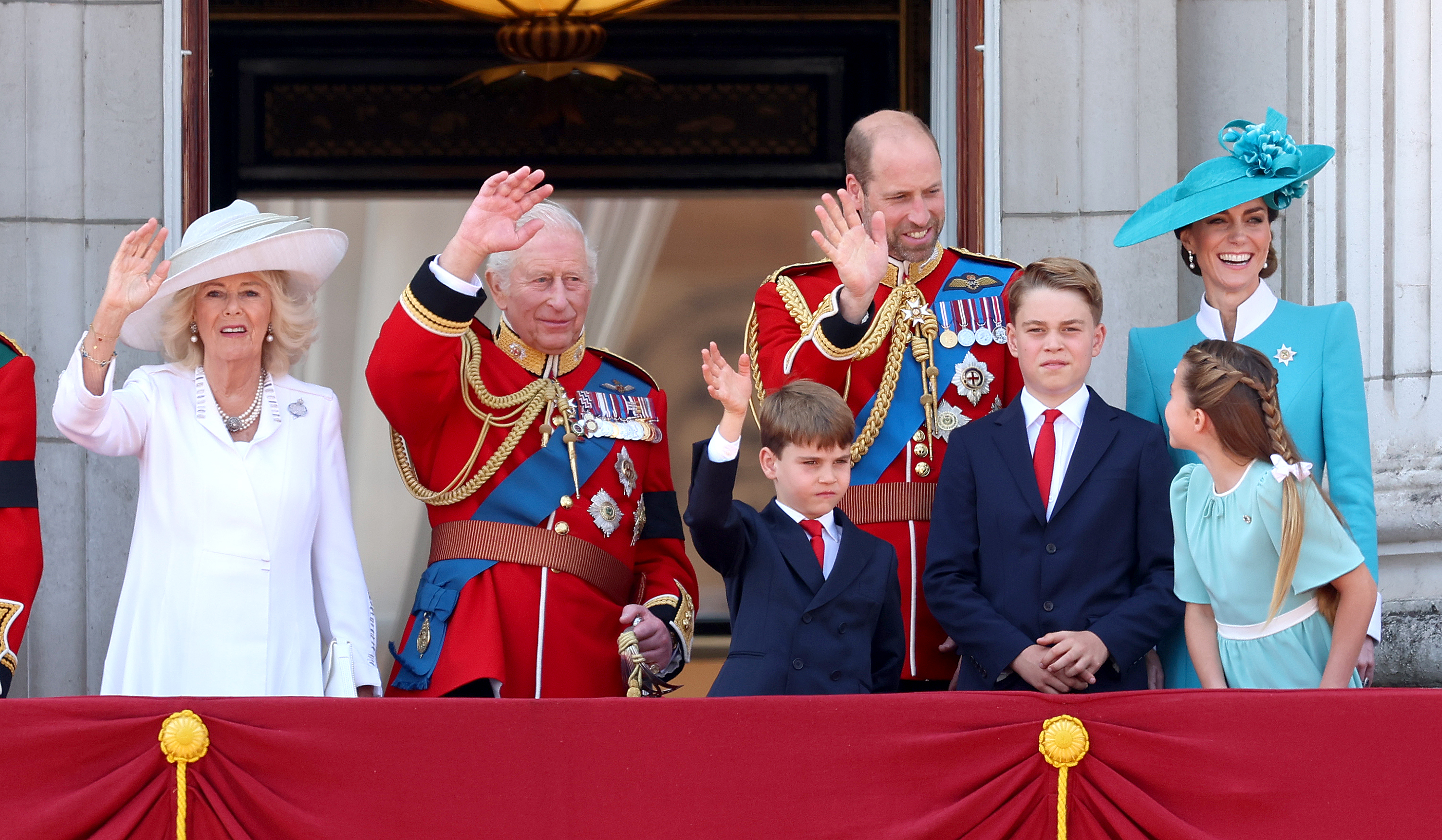 Königin Camilla, König Charles III., Prinz Louis, Prinz William, Prinz George, Prinzessin Charlotte und Prinzessin Catherine grüßen die Menge während der Trooping the Colour-Zeremonie 2025. Prinzessin Charlotte blickt zu ihrem Vater zurück, Prinz George posiert mit gekreuzten Handgelenken, Prinzessin Catherine lächelt und die anderen Mitglieder der königlichen Familie winken der Menge zu.