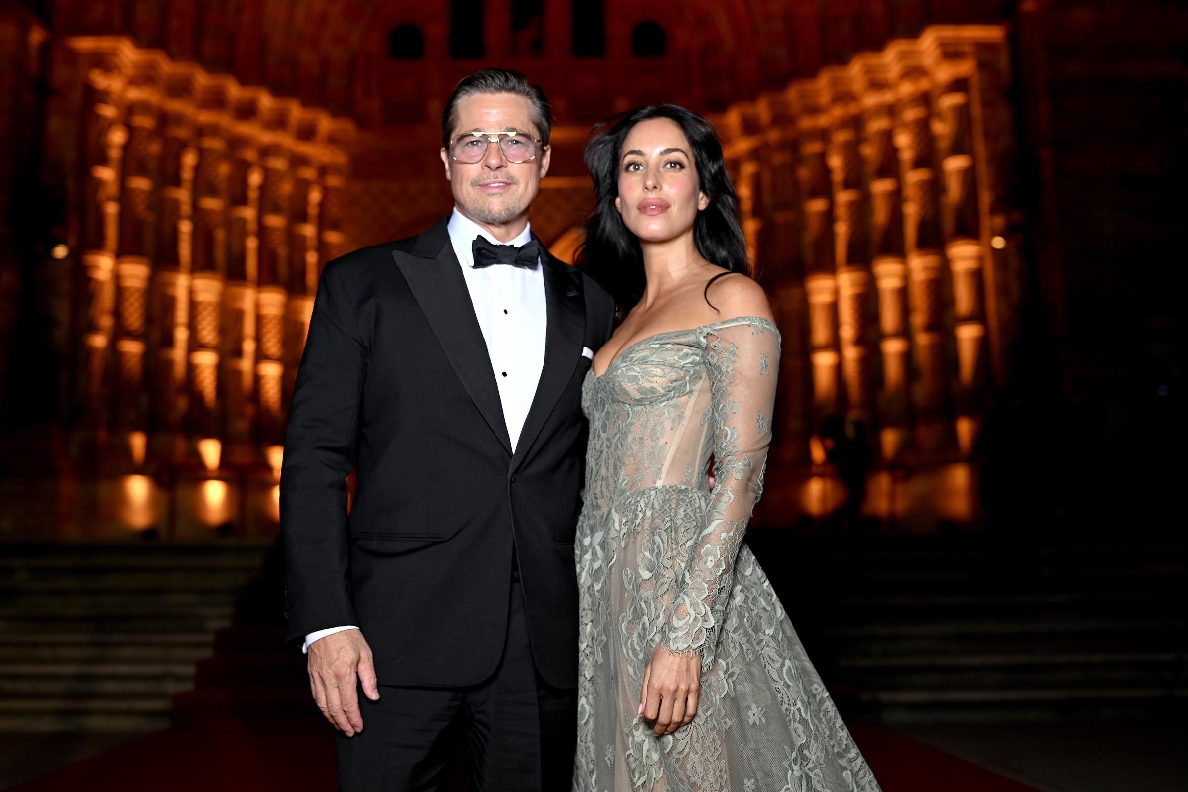 Brad Pitt und Ines de Ramon posieren gemeinsam auf der beleuchteten Treppe im „2026 Albies“ in London – Brad in einem schwarzen Smoking und mit getönten Sonnenbrillen, Ines in einem transparenten, schulterfreien Spitzenkleid mit aufwendigen Blumenstickereien, einem eng anliegenden Korsett und einer wallenden Schleppe. Die zarten Details des Kleides und die durchscheinenden Ärmel heben sich deutlich von der warmen, goldenen Beleuchtung des Veranstaltungsortes ab. | Quelle: Getty Images