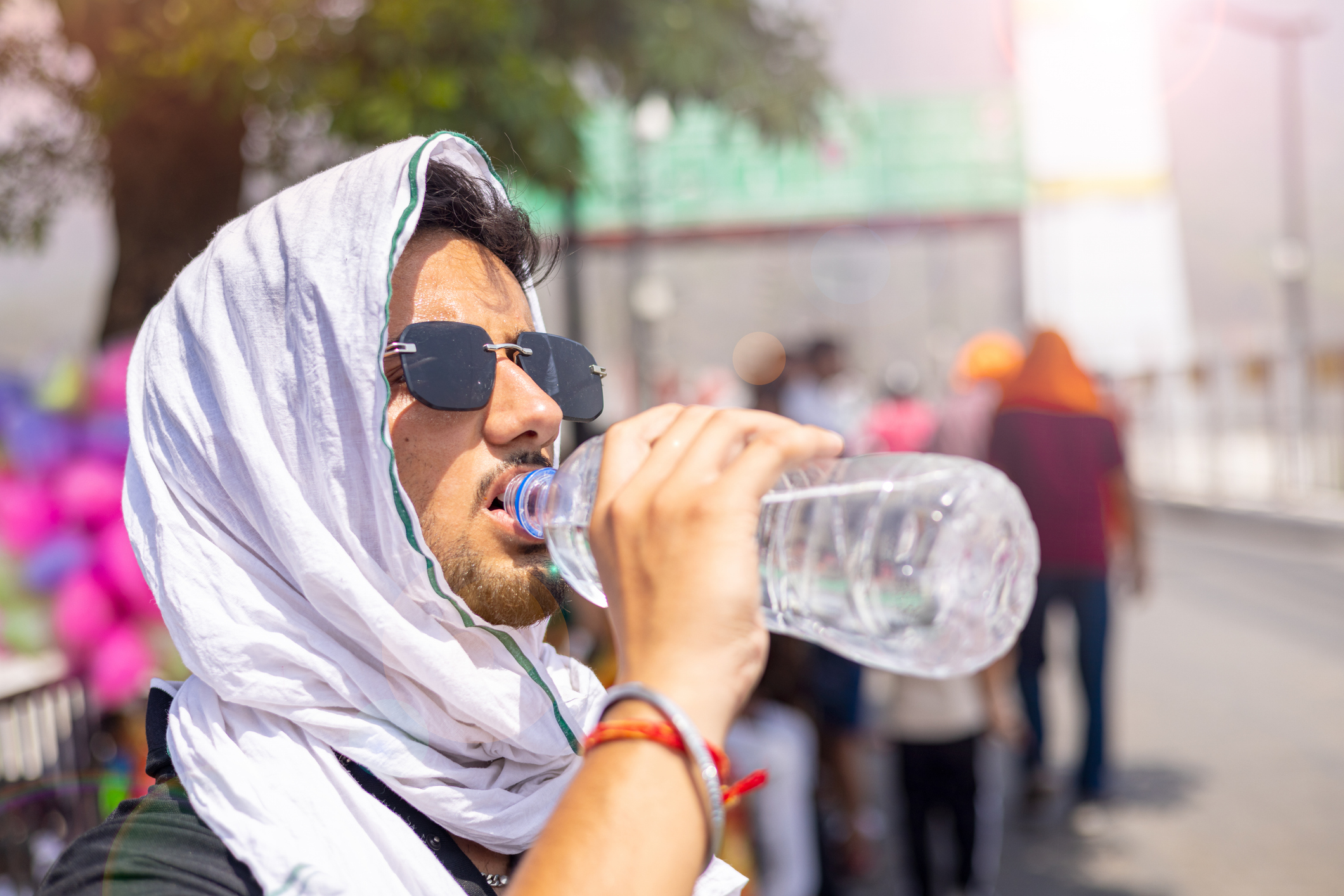 Nahaufnahme eines jungen Mannes, der sich an einem extrem heißen Nachmittag den Kopf mit einem Tuch bedeckt und eine schwarze Sonnenbrille trägt, während er Wasser aus einer Plastikflasche trinkt. | Quelle: Getty Images
