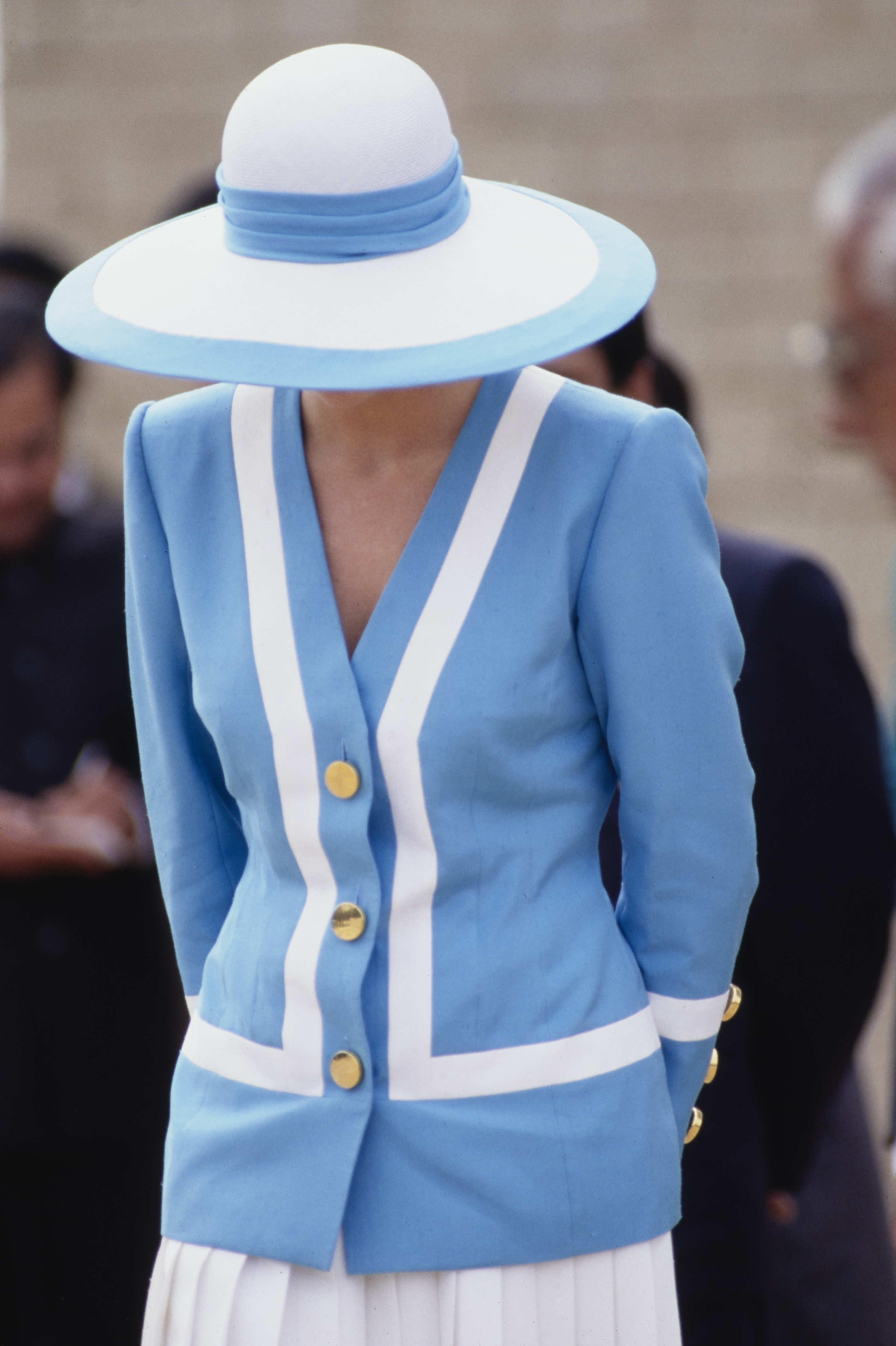 Prinzessin Diana steht mit ruhiger Gelassenheit auf dem Delhi War Cemetery, gekleidet in ihrem charakteristischen blau-weißen Anzug und einem breitkrempigen Hut. Das Bild fängt ihre feierliche Anmut inmitten der ruhigen Umgebung ein, ein Spiegelbild des respektvollen Gedenkens.