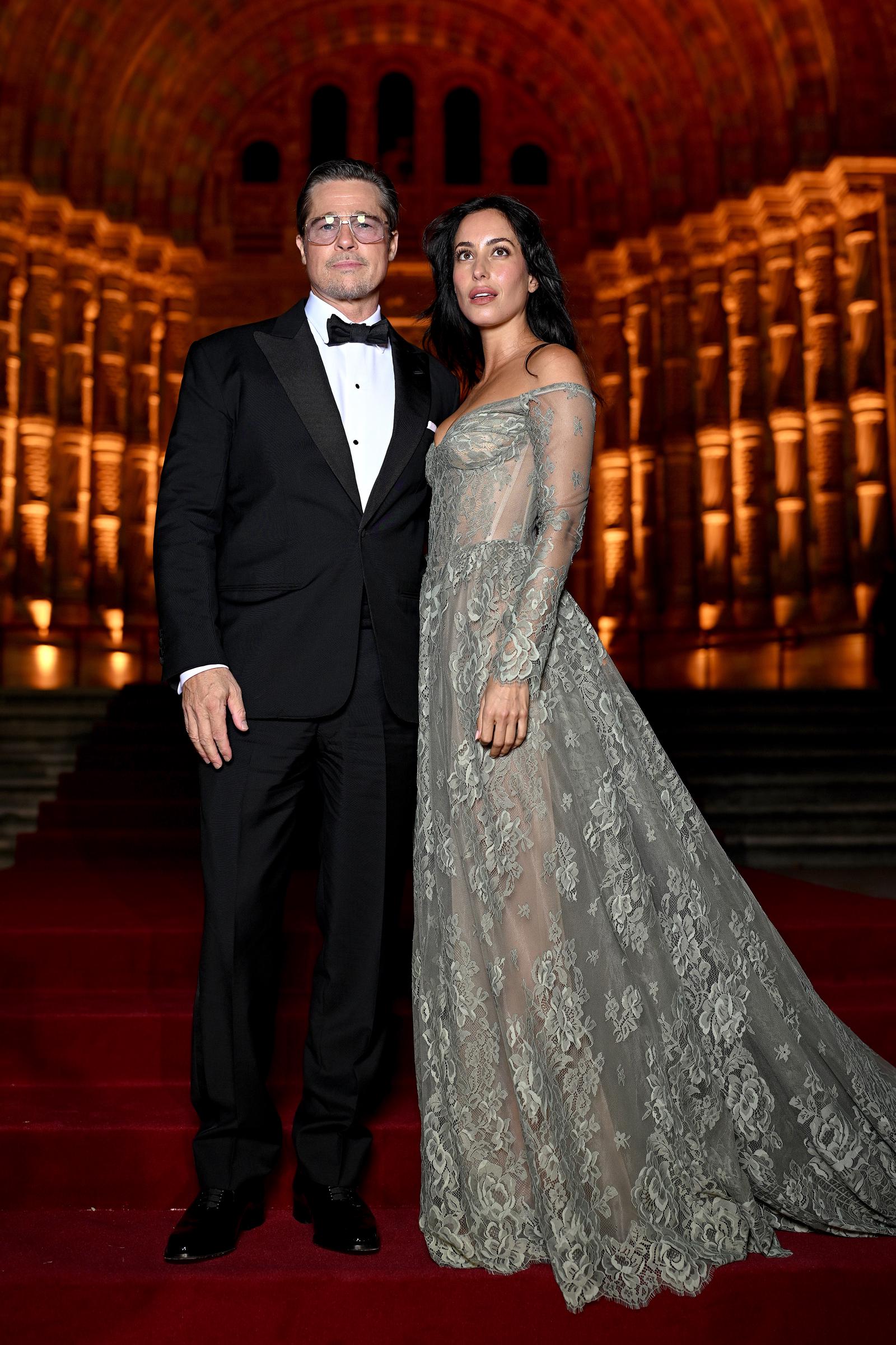 Brad Pitt und Ines de Ramon posieren auf der Treppe vor dem roten Teppich für ein Ganzkörperfoto und präsentieren dabei ihre kontrastreichen Abendoutfits. Ines’ bodenlanges, besticktes Abendkleid hat eine dramatische Schleppe, die sich über die Stufen ausbreitet, während die leuchtend goldenen Bögen des Veranstaltungsortes eine beeindruckende Kulisse bilden. | Quelle: Getty Images