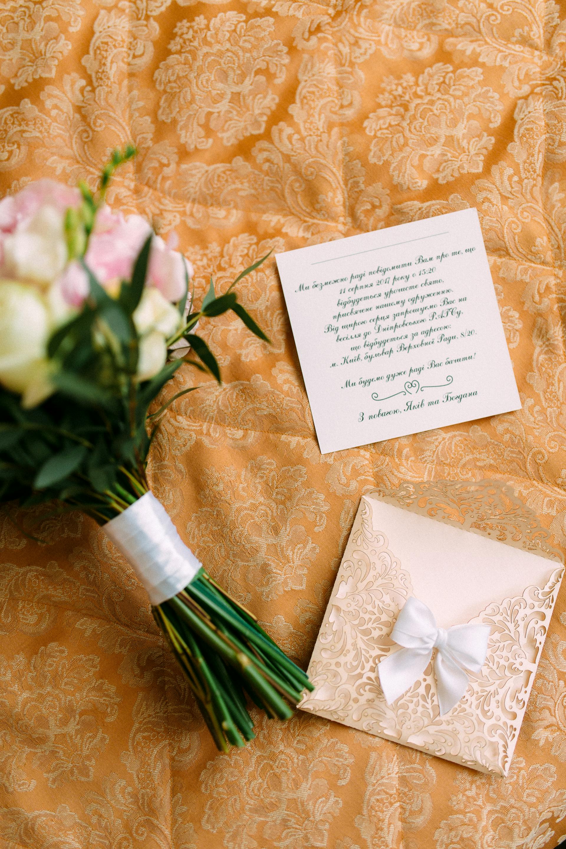 Eine Hochzeitseinladung, ein Umschlag mit einer Schleife und ein Blumenstrauß auf einem goldenen, gemusterten Stoff | Quelle: Pexels