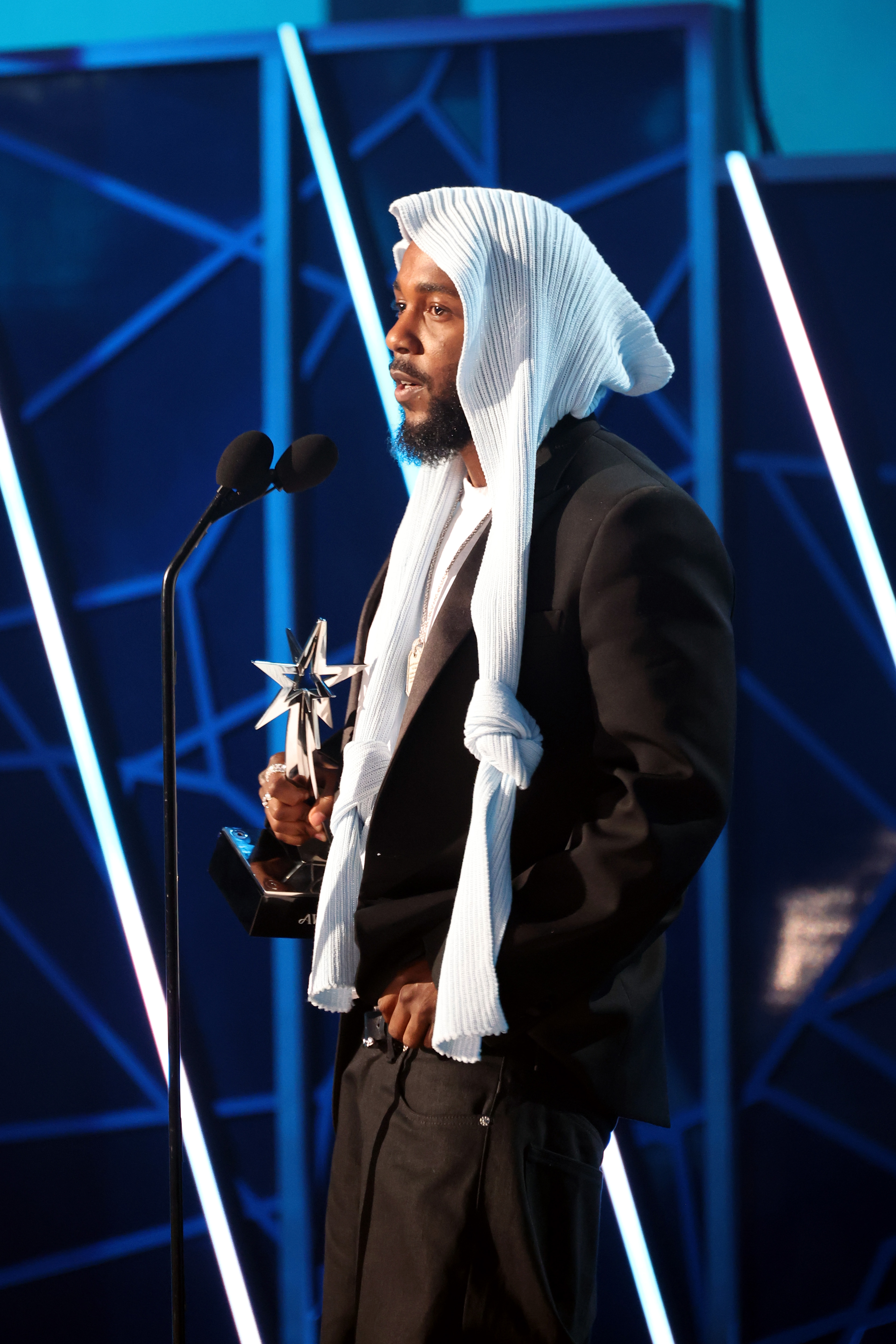 Mit einer ruhigen und doch kraftvollen Präsenz wendet sich Kendrick Lamar an das Publikum, nachdem er bei den BET Awards 2025 gewonnen hat. Gekleidet in ein schwarz-weißes Ensemble, hielt der Künstler seine Trophäe stolz in die Höhe, während er vor der leuchtenden geometrischen Kulisse stand.