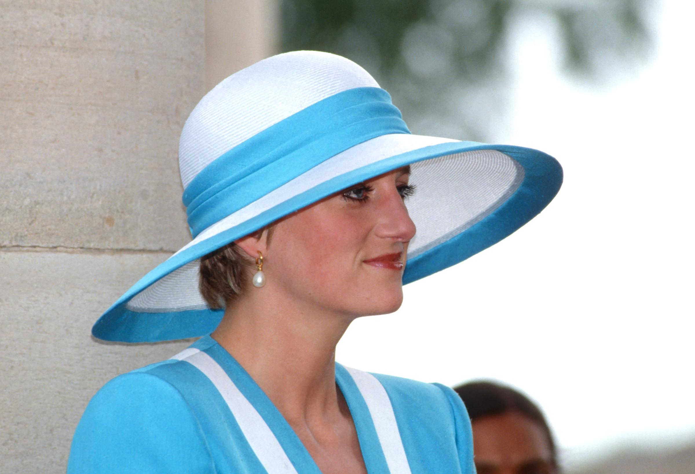 In einer Nahaufnahme von ihrem Besuch auf dem Delhi War Cemetery im Jahr 1992 steht Prinzessin Dianas gelassener Gesichtsausdruck im Mittelpunkt. In einem eleganten blau-weißen Anzug von Catherine Walker und einem formschönen Hut von Philip Somerville fängt die Kamera die ruhige Würde in ihrem Gesicht ein, das von ihrem unverkennbaren Stil elegant eingerahmt wird.