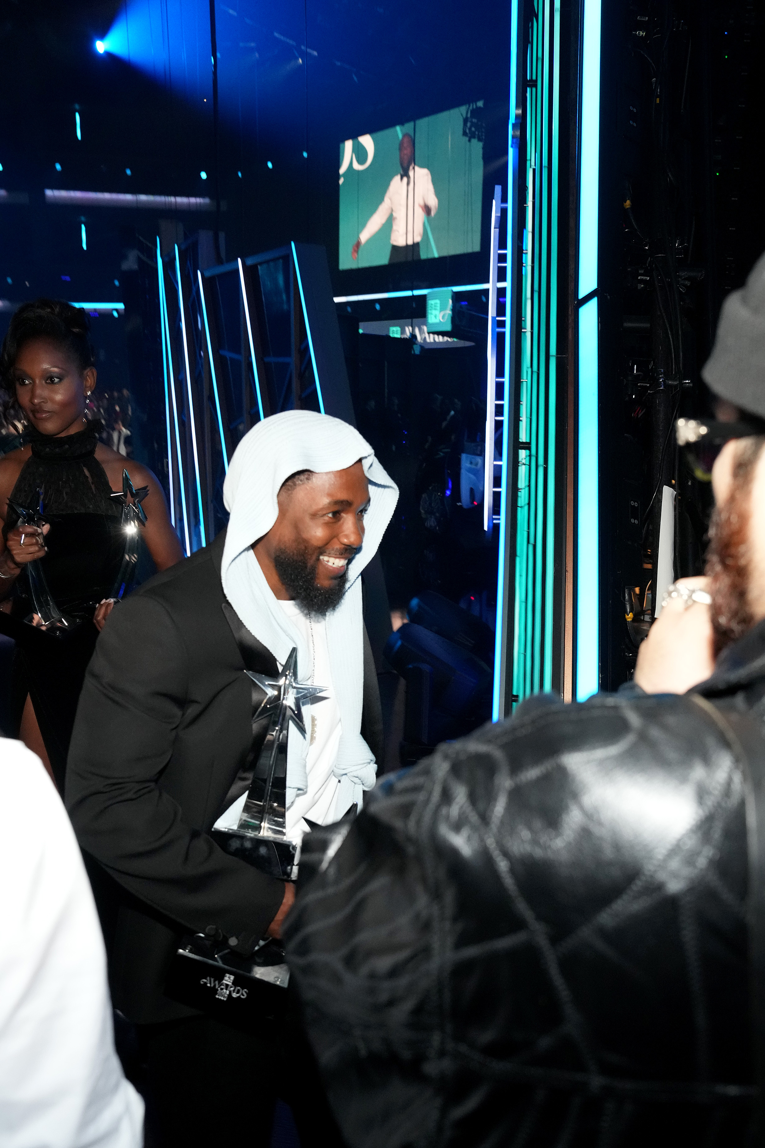 Hinter der Bühne des Peacock Theaters lächelt Kendrick Lamar, als er nach seinem Sieg bei den BET Awards 2025 Gratulanten begrüßt und mehrere Preise in die Hand nimmt. Zu seinem mehrschichtigen Look gehörte ein gestricktes Kopfteil, das vorne zusammengebunden wurde.