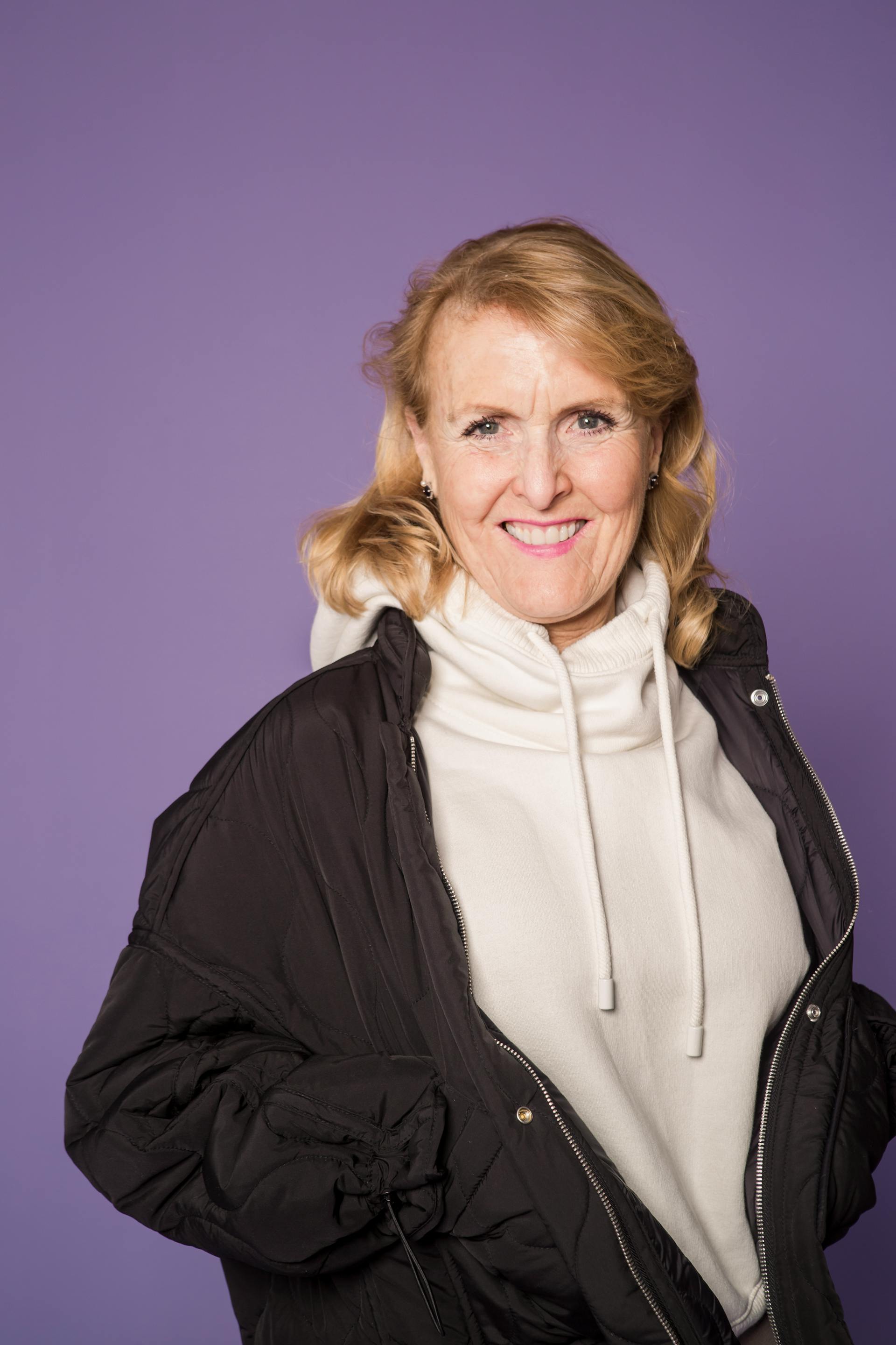 Eine glückliche reife Frau in einer schwarzen Jacke und einem weißen Hoodie lächelt vor einem lila Hintergrund | Quelle: Pexels
