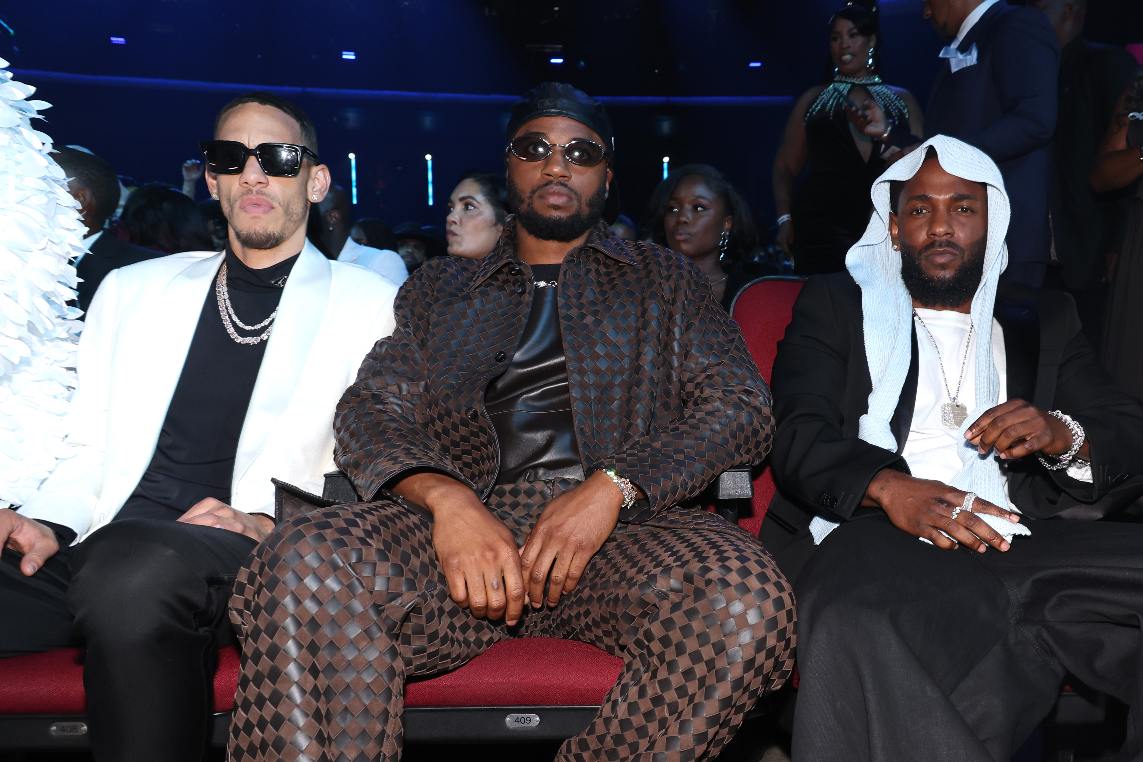 Bei den BET Awards 2025 sitzt Kendrick Lamar in der ersten Reihe und posiert mit seinen Kollegen King Matta und Dave Free in einem ruhigen, entspannten Moment. Er trug sein mittlerweile bekanntes Headpiece zusammen mit einem eleganten schwarzen Anzug und einem weißen Hemd.