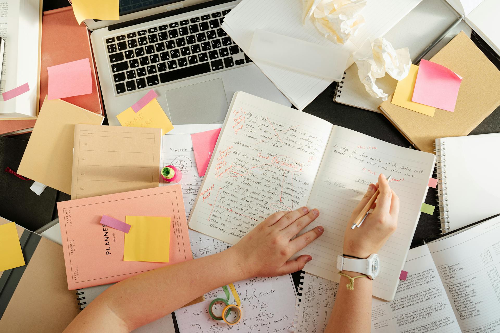 Ein unordentlicher Schreibtisch mit Büchern, einem Laptop und einer Person, die in ein Notizbuch schreibt | Quelle: Pexels