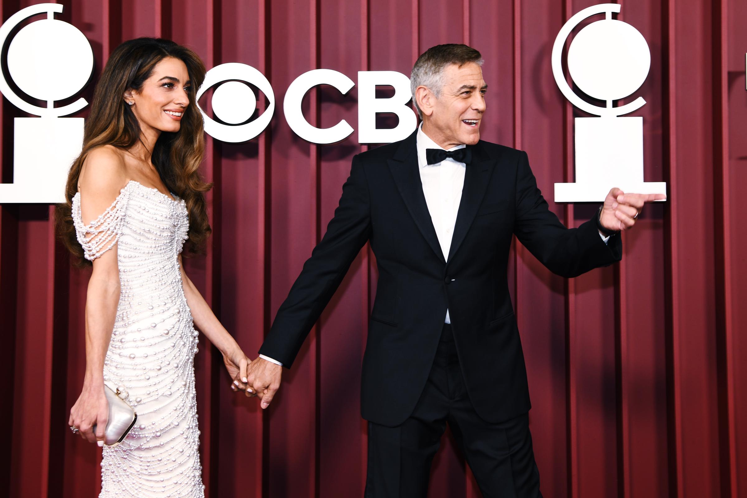 Amal Clooney strahlt, während George Clooney ausdrucksvoll plaudert und ihre Finger auf dem roten Teppich ineinander verschränkt. Ihr perlenbesetztes Kleid schimmert im Licht, während er die Stimmung mit einer verspielten Geste aufrecht erhält.