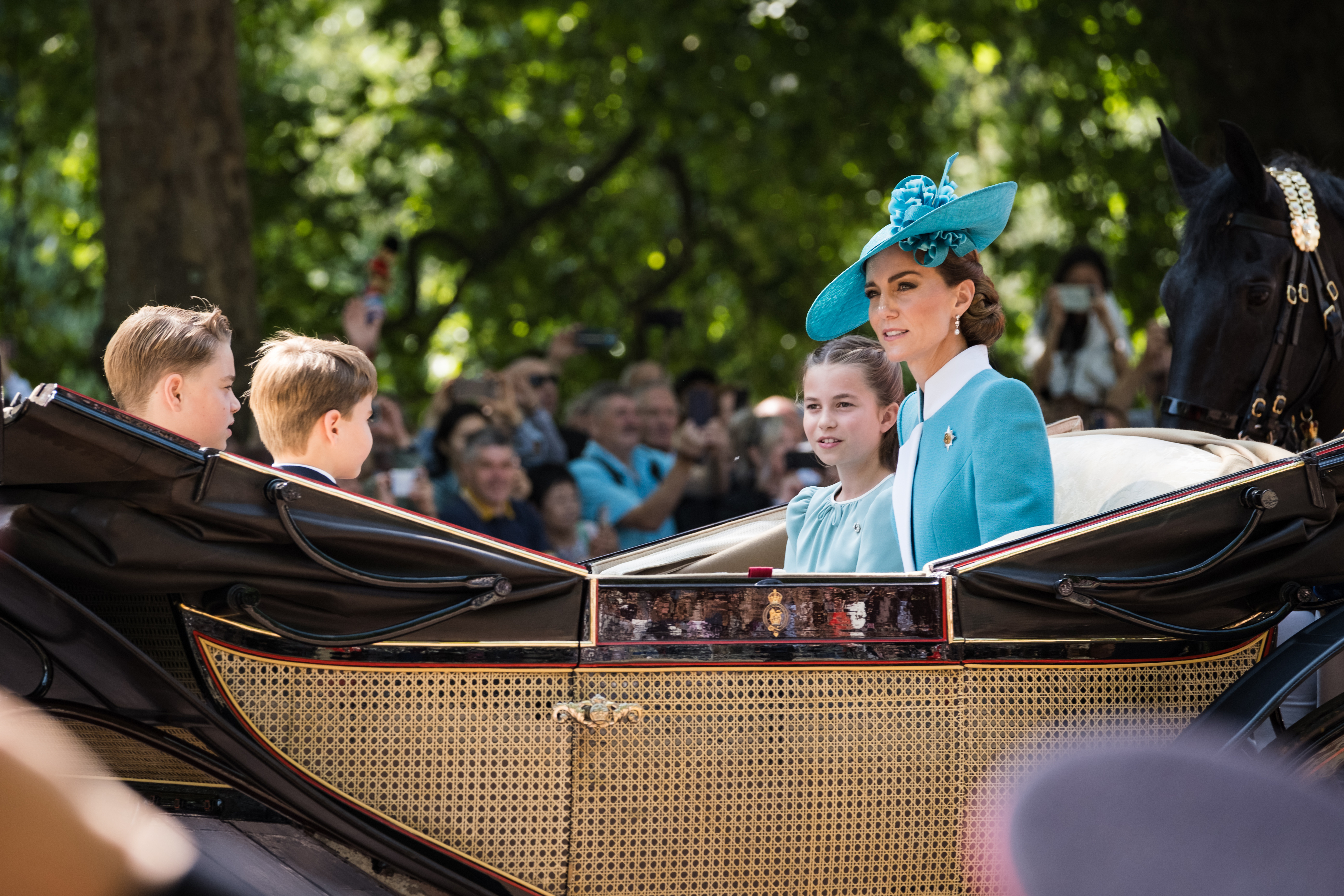 Prinzessin Charlotte, Prinzessin Catherine, Prinz George und Prinz Louis fahren in der königlichen Kutsche, während die Leute zuschauen und Fotos von der Mutter mit ihren Kindern machen.