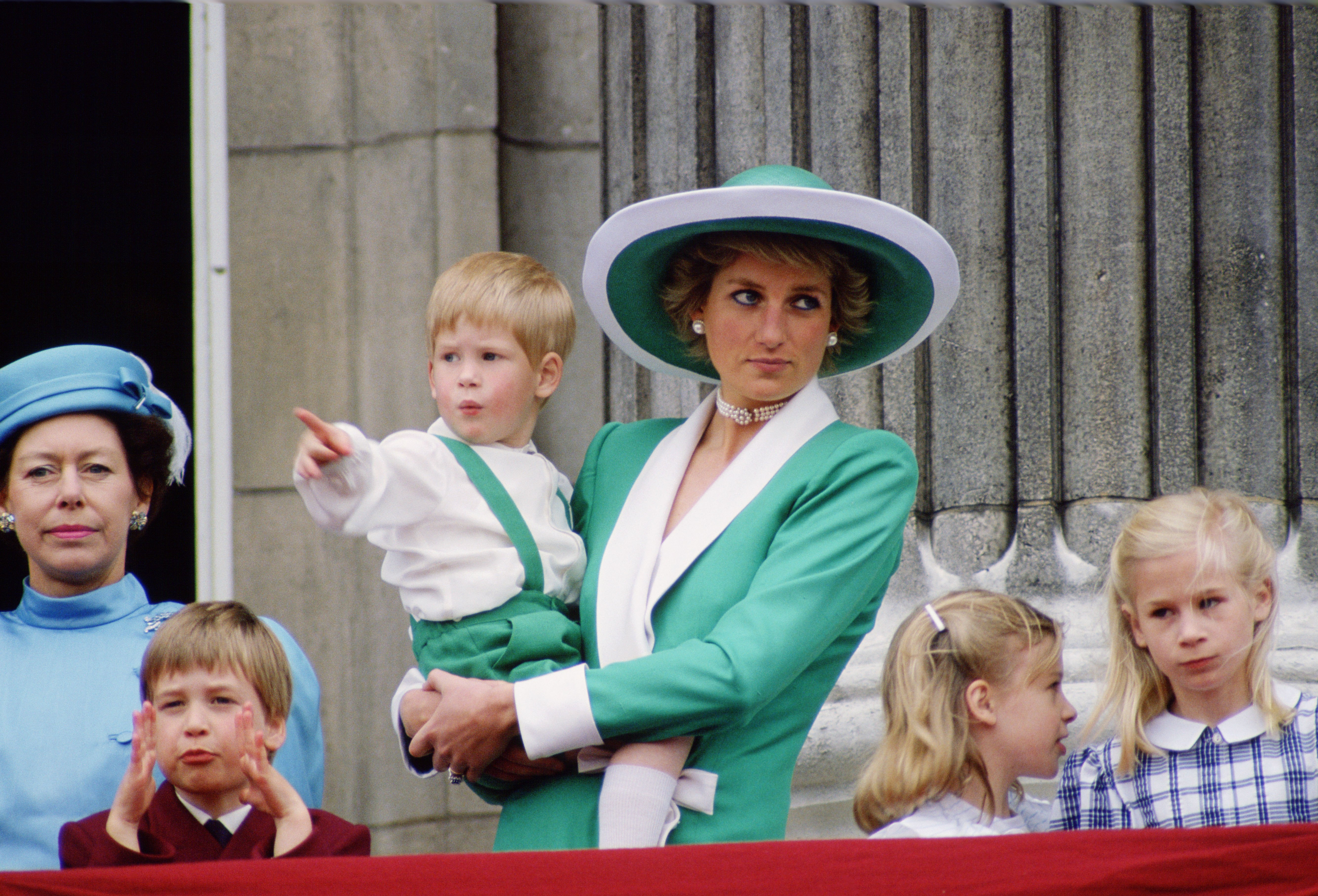 Prinzessin Diana, gekleidet in einen grün-weißen Anzug mit passendem Hut, hält den jungen Prinz Harry im Arm, während sie vom Balkon des Buckingham Palastes aus das Trooping the Colour 1988 beobachtet. Umgeben von Prinz William, Lady Rose Windsor, Lady Davina Windsor und Prinzessin Margaret fängt Dianas ruhige Präsenz einen zeitlosen Moment der königlichen Familie ein, der von Tradition und Prunk umrahmt ist.