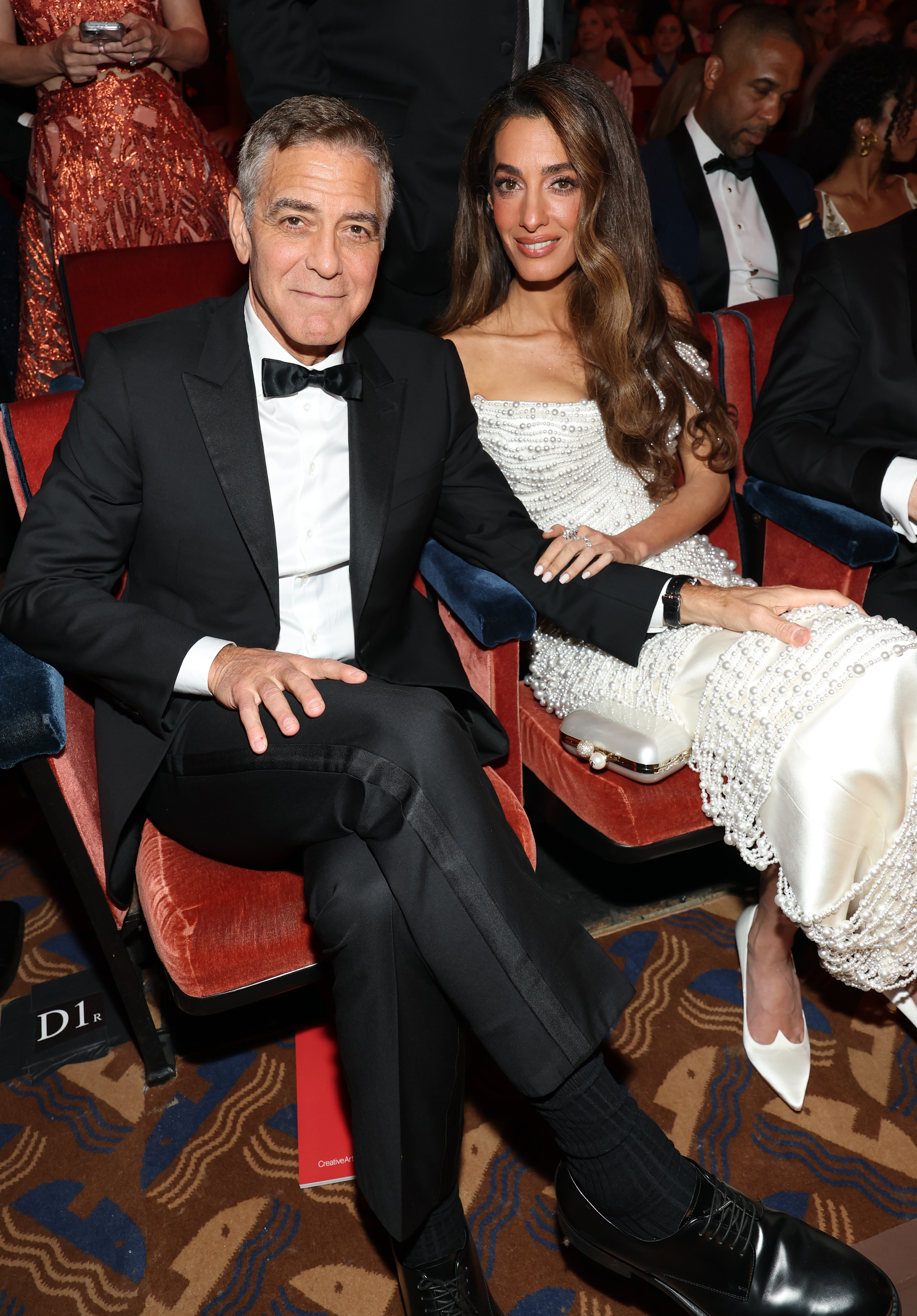 Im Saal sitzt George Clooney mit gekreuzten Beinen, während Amal sich an ihn lehnt und eine Hand auf seinem Arm ruht. Das Paar nimmt eine entspannte Pose ein, umgeben von anderen Gästen, während sie auf den Beginn der Zeremonie warten.