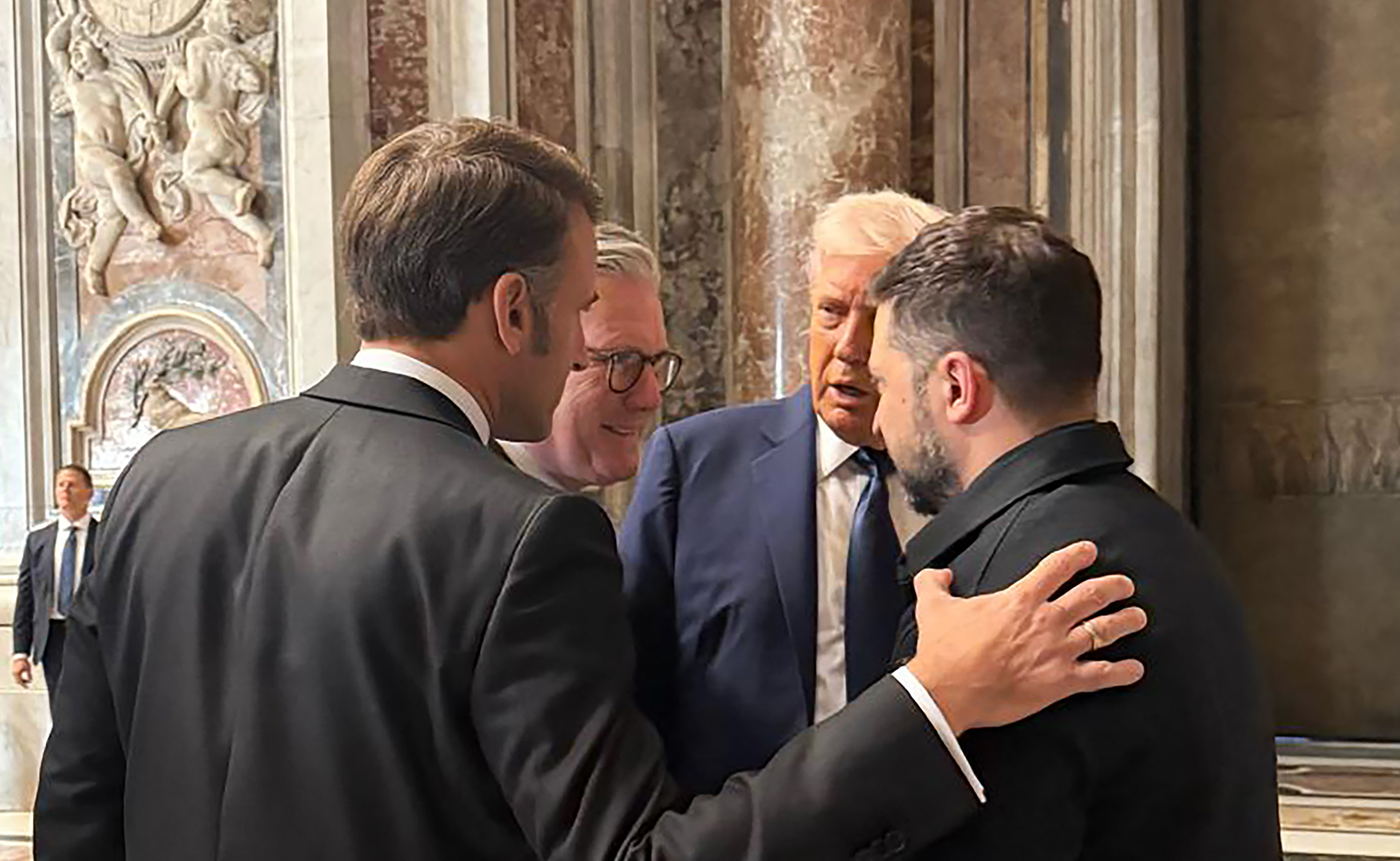Bei der Beerdigung von Papst Franziskus legt der französische Präsident Emmanuel Macron dem ukrainischen Präsidenten Wolodymyr Zelenskij tröstend die Hand auf die Schulter und bietet ihm seine Unterstützung inmitten des anhaltenden Krieges an. Neben ihnen stehen der britische Premierminister Keir Starmer und Donald, während sie sich unterhalten.
