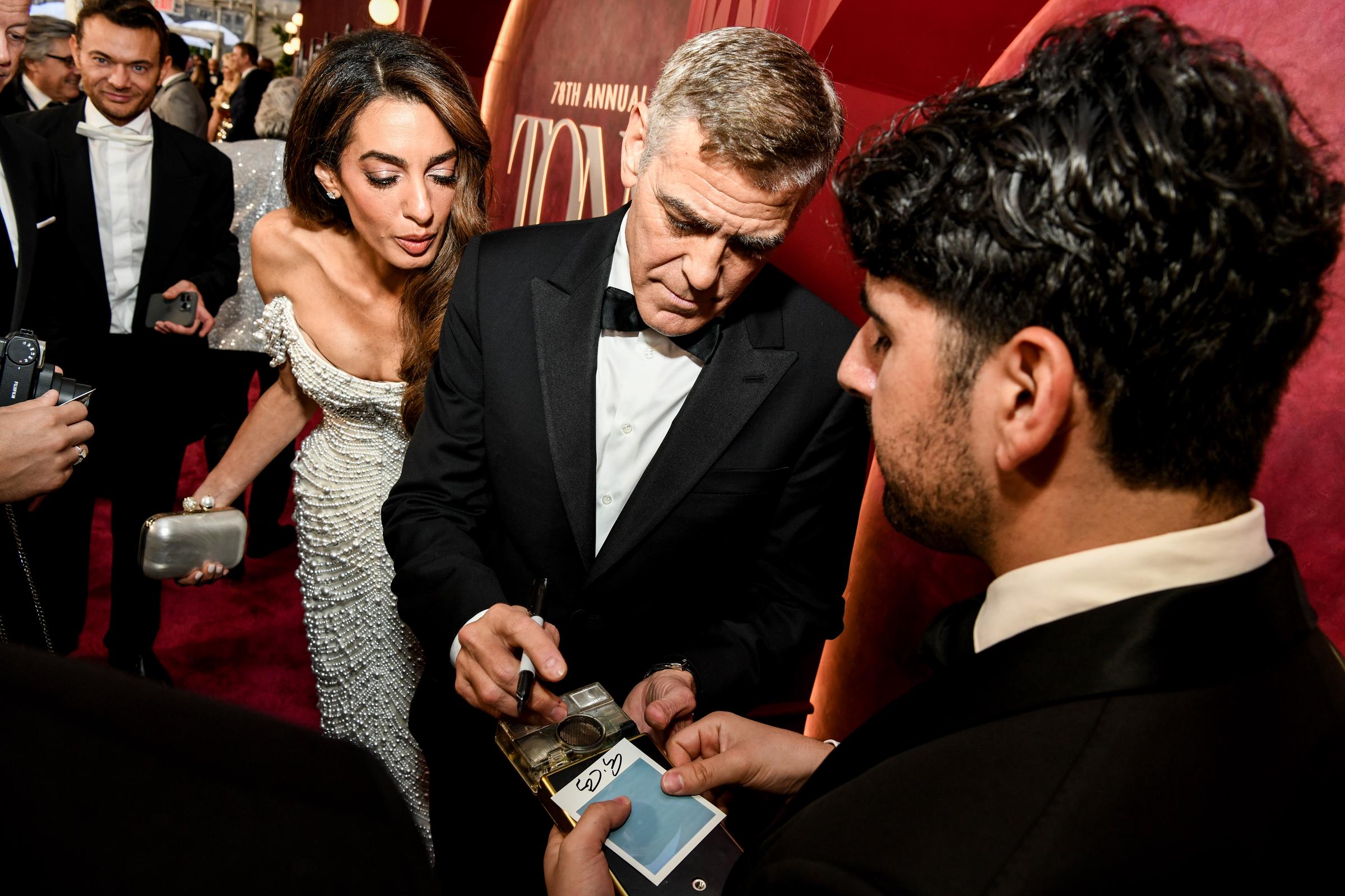 George Clooney signiert Erinnerungsstücke, während Amal Clooney sich vorbeugt und mit konzentrierter Miene beobachtet. Das Paar hält inmitten eines Wirbels von Fans und blitzenden Kameras auf dem roten Teppich inne.