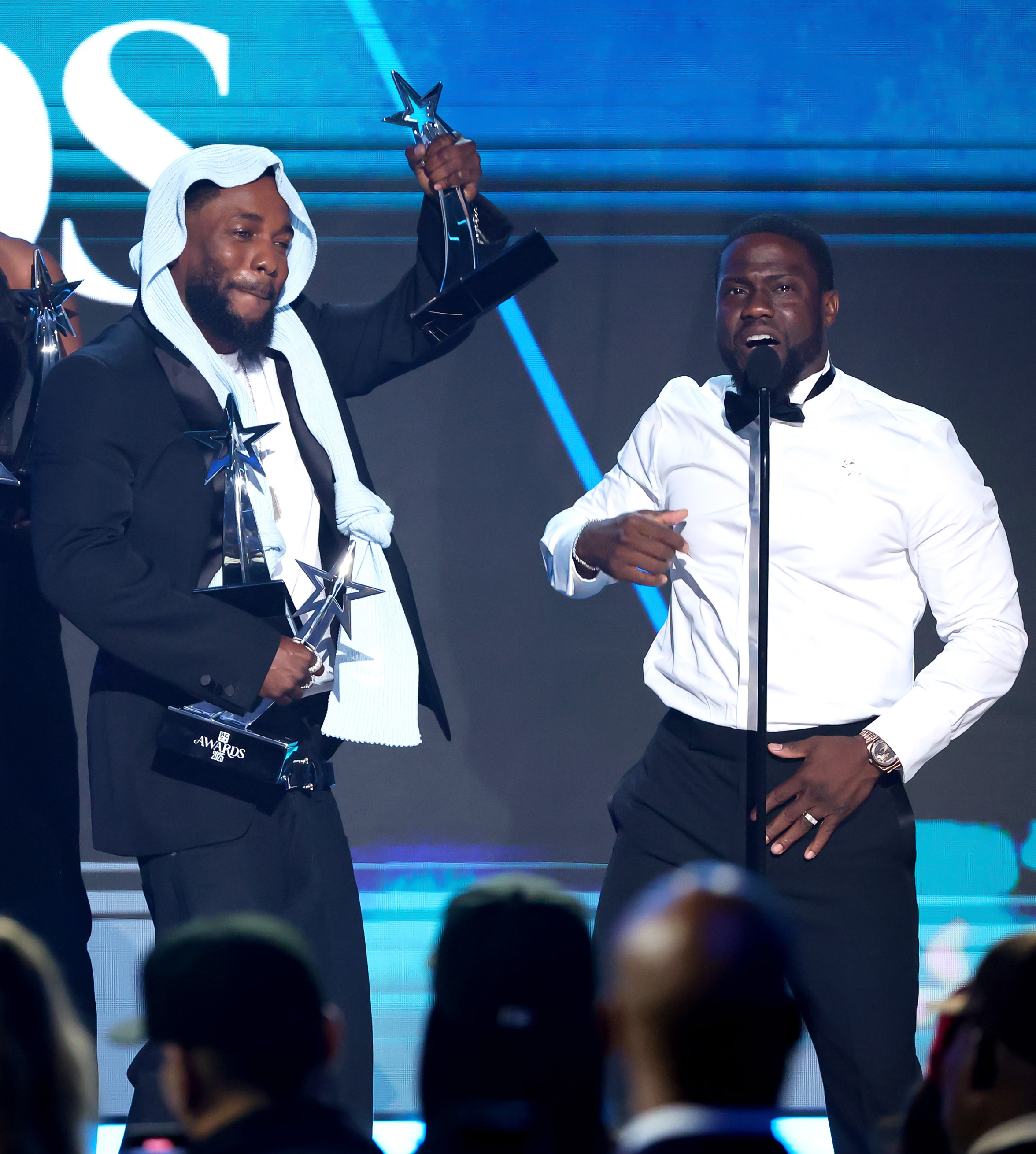 Mit einer seiner Trophäen in der Hand steht Kendrick Lamar neben Kevin Hart, als sie bei den BET Awards 2025 einen heiteren Moment teilen. Lamars mehrfache Siege wurden zu einem Running Gag, als Hart versuchte, ihm beim Tragen der Last zu helfen.