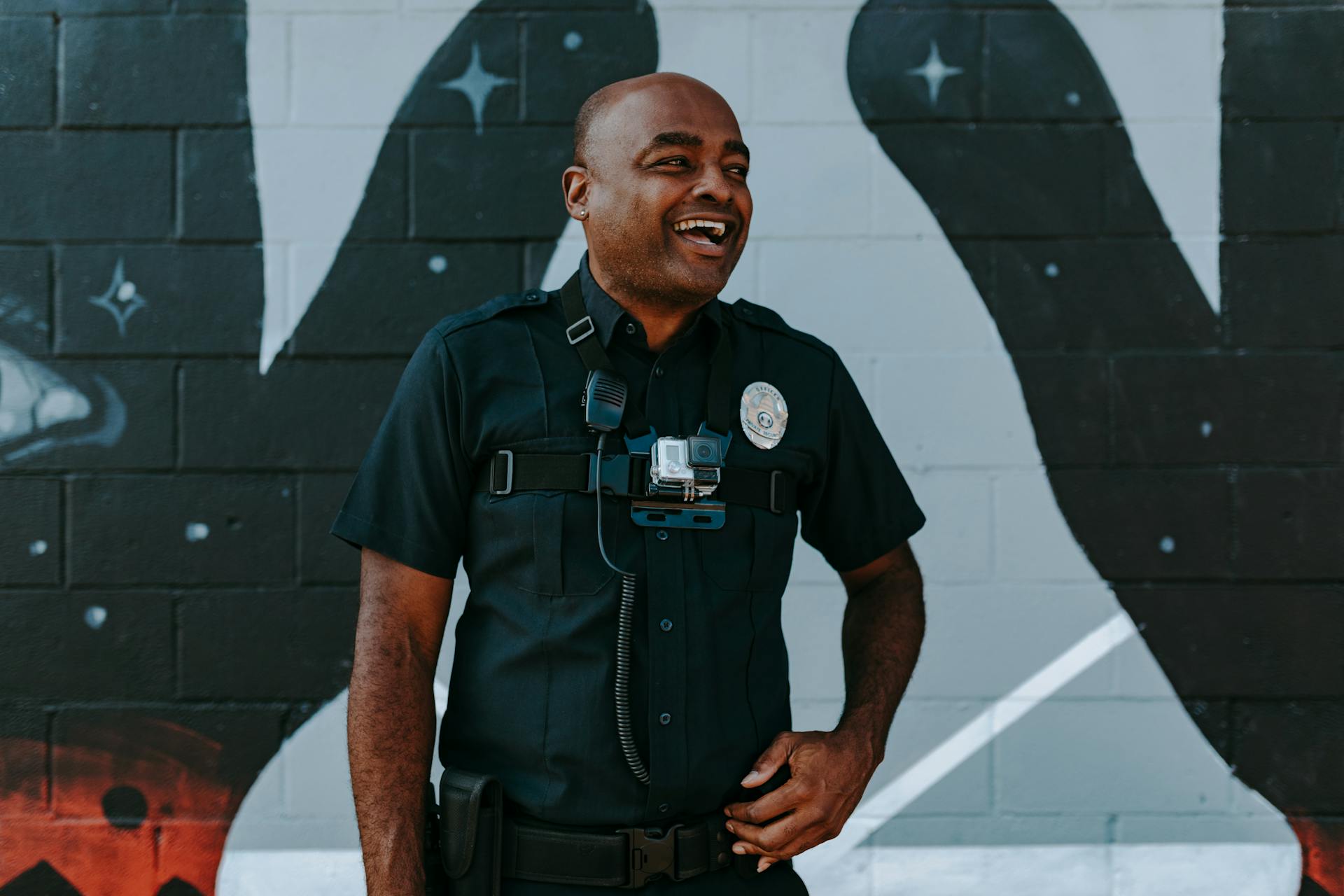 Ein Polizist mit einer Körperkamera lacht vor einer Graffiti-Wand | Quelle: Pexels