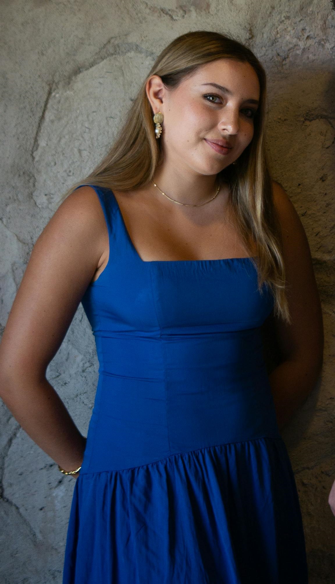 Eine lächelnde junge Frau in einem blauen Kleid mit einer Halskette und Ohrringen steht vor einer Steinmauer | Quelle: Pexels