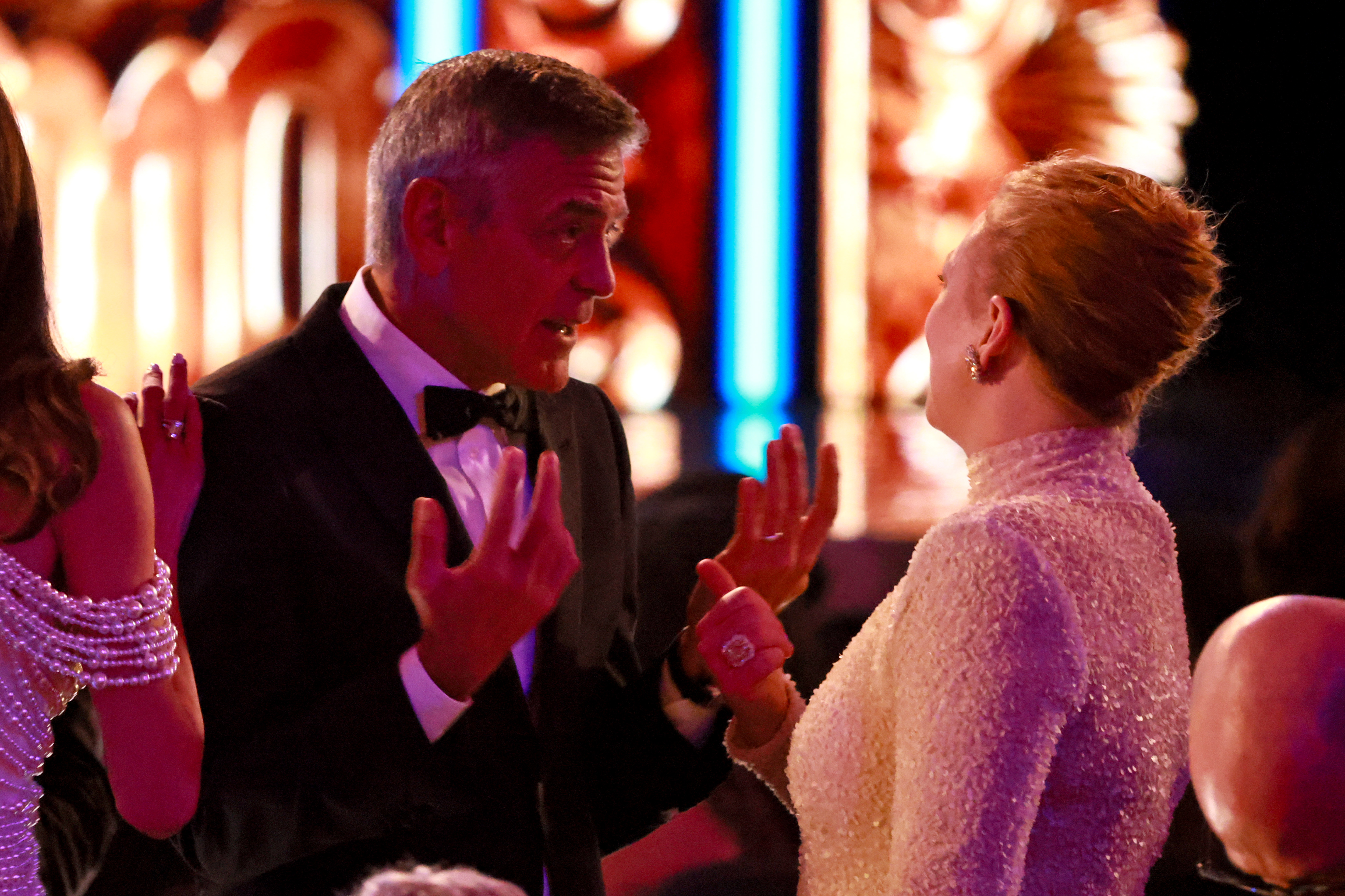 Mit ausdrucksstarken Gesten plaudert George Clooney mit Sarah Snook mitten in der Vorstellung, während Amal Clooney neben ihm steht. Der lebhafte Moment entfaltet sich in dem warm beleuchteten Theater.